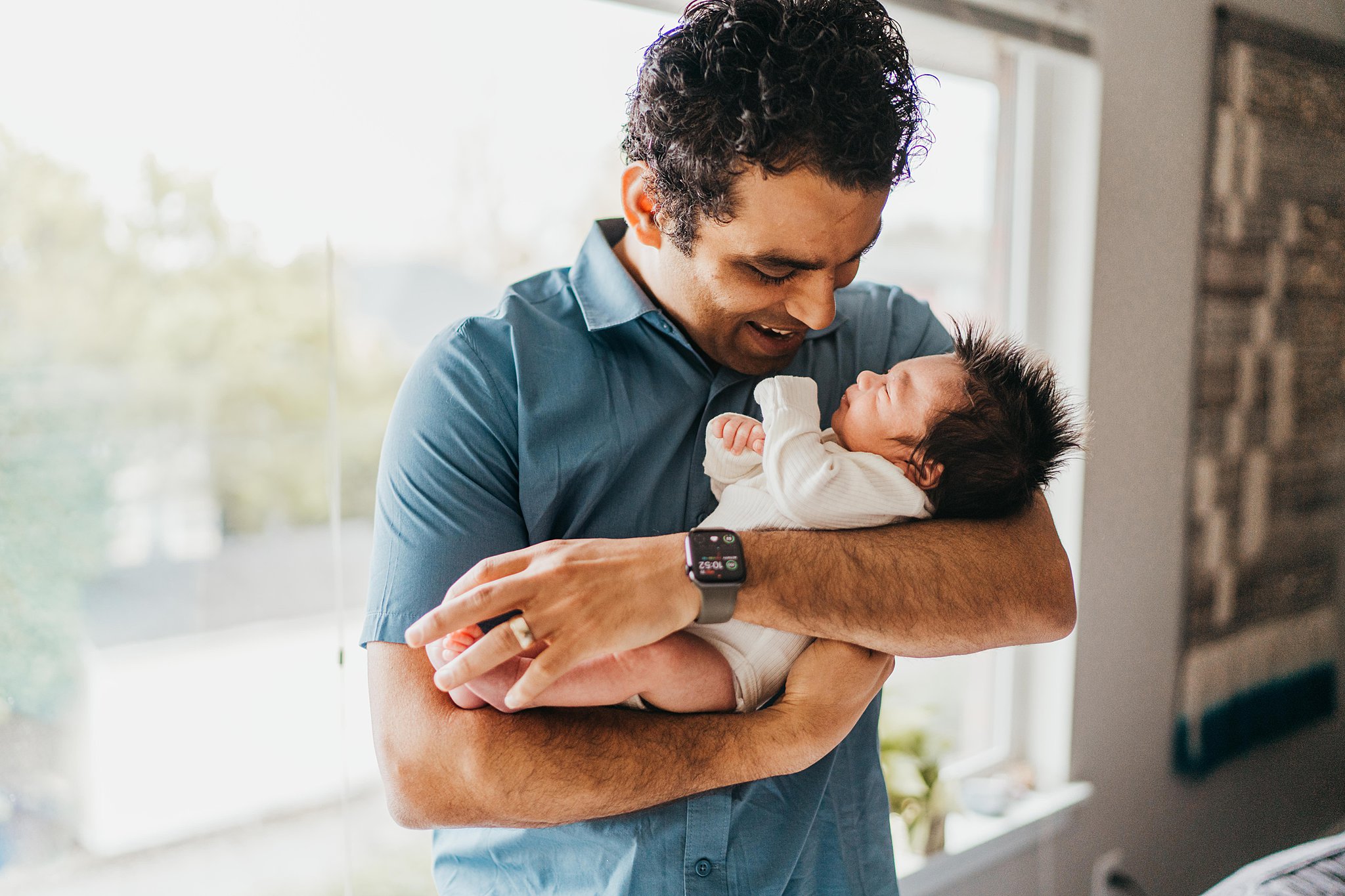 A new father smiles down at his sleeping newborn baby in his arms while standing by a window baby diaper service seattle