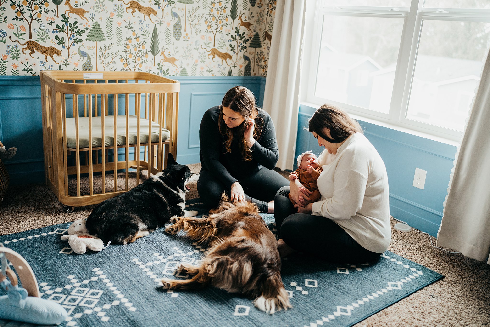 New moms sit on the floor of their nursery while playing with their two dogs and their newborn baby center for women's health at evergreen