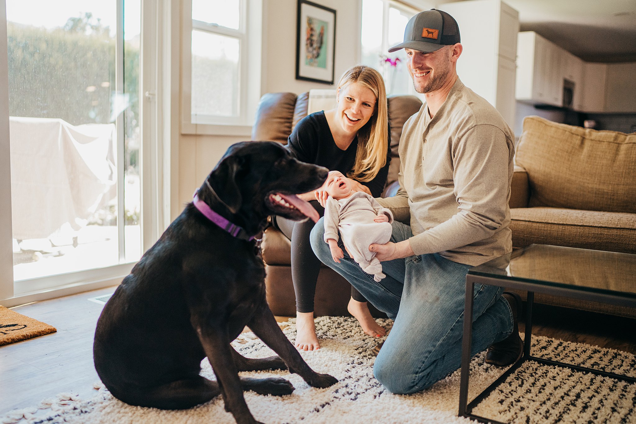 A new father shows his newborn baby to the family black lab while mom sits on a leather chair in living room fit4mom eastside