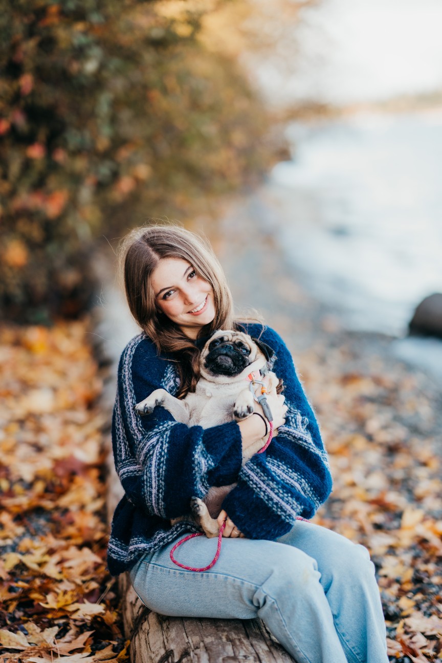 a portrait of a high school girl holding her dog sitting on a log in front of beautiful foliage at luther burbank park