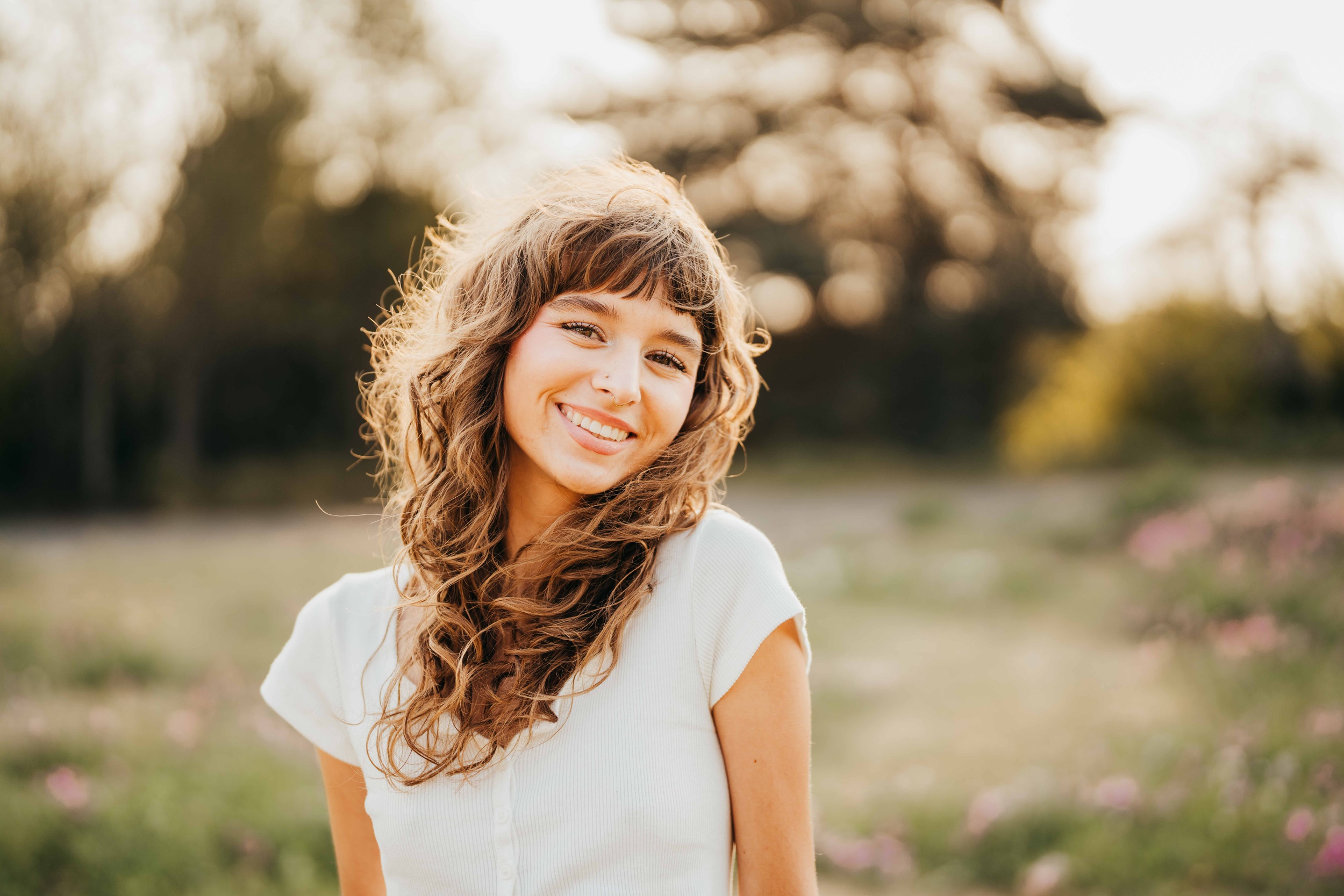 A high school senior in a white shirt smiles while exploring a field of pink flowers