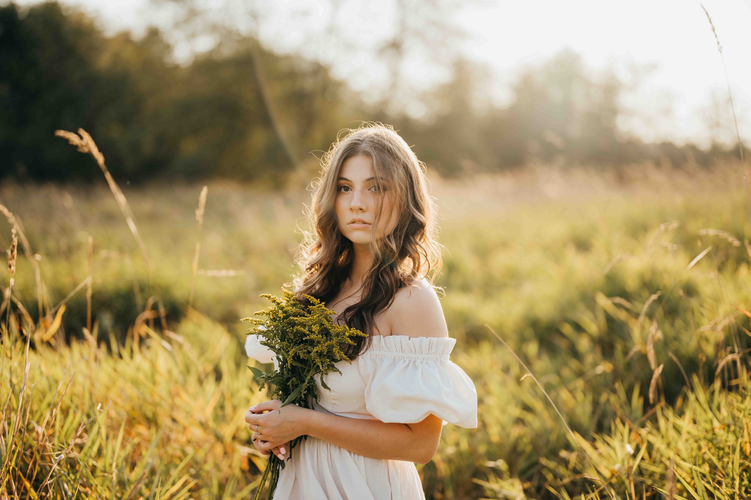 A teenage girl in a white dress walks in a field of tall grass holding wildflowers