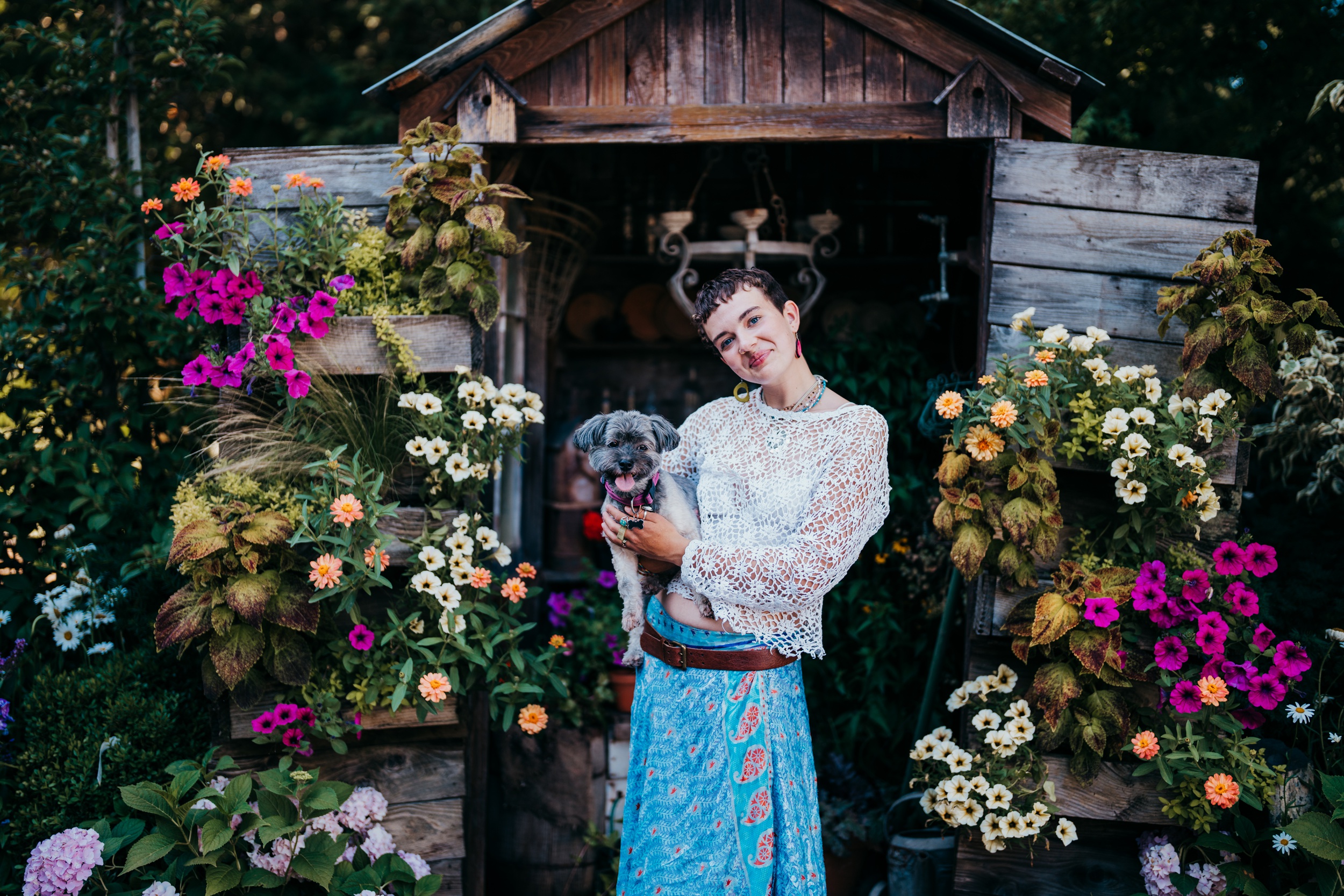 A happy Bellevue High School senior in a lace top and blue skirt stands with their dog by a garden shed