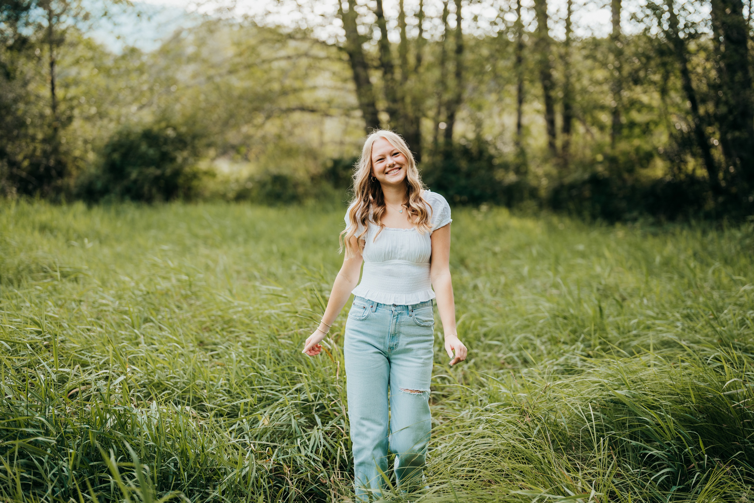 A smiling teenage girl in a blue blouse and jeans walks in a meadow of tall green grass