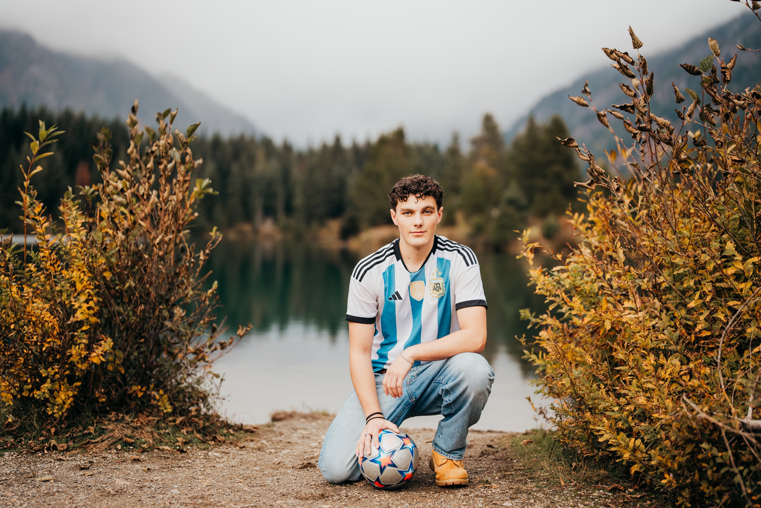 A teenage soccer player kneels with a ball by a lake in the mountains