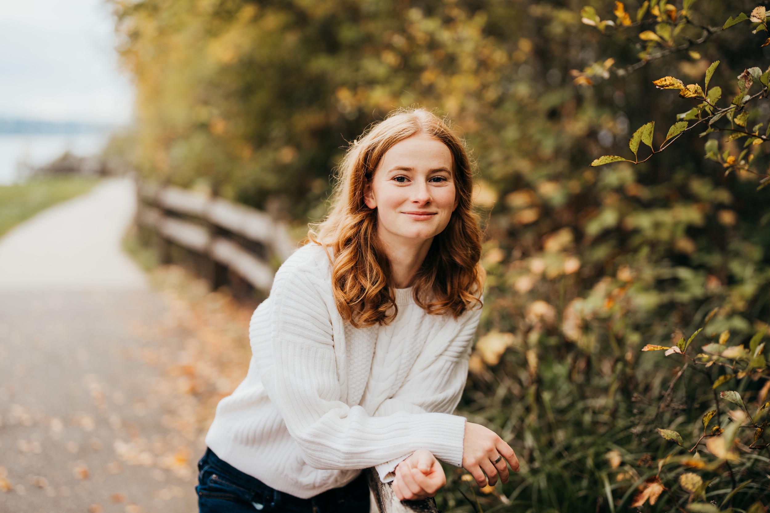 A Newport High School senior in a white sweater leans on a park fence smiling