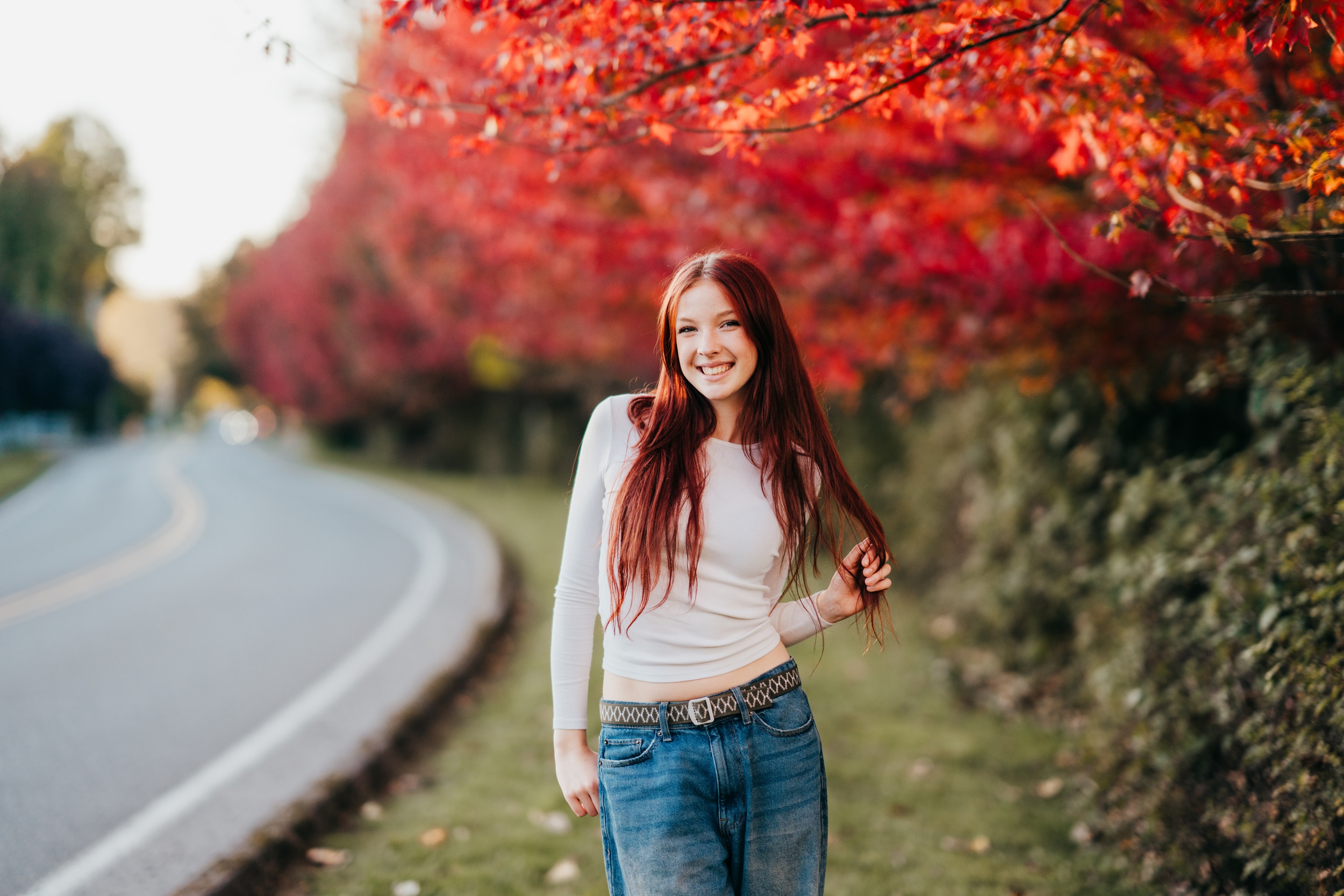 A teenage girl in jeans and white shirt smiles while walking by the road under red leafed trees at sunset