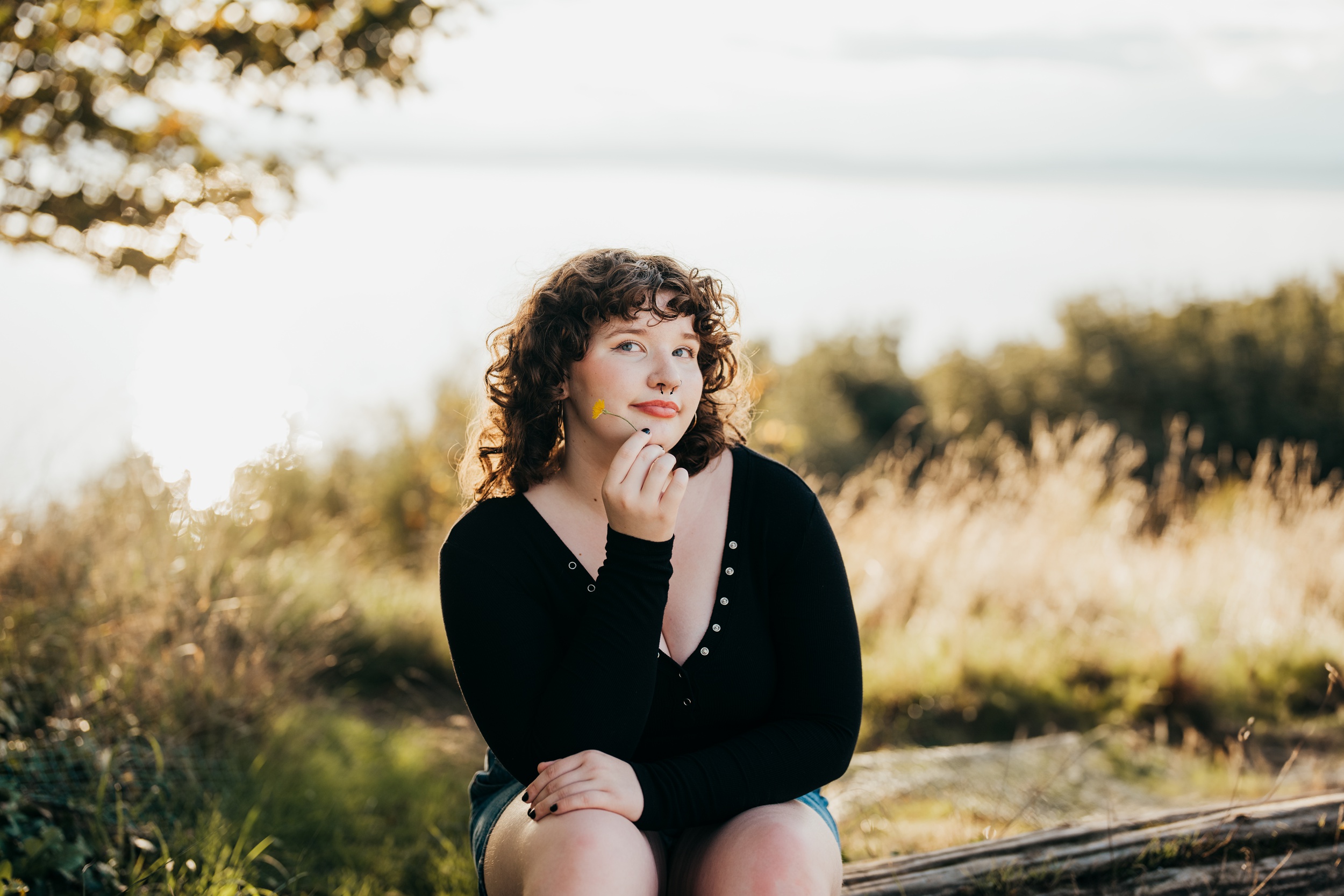 A Roosevelt High School senior sits on a log in a park holding a yellow wildflower by her cheek in a black blouse