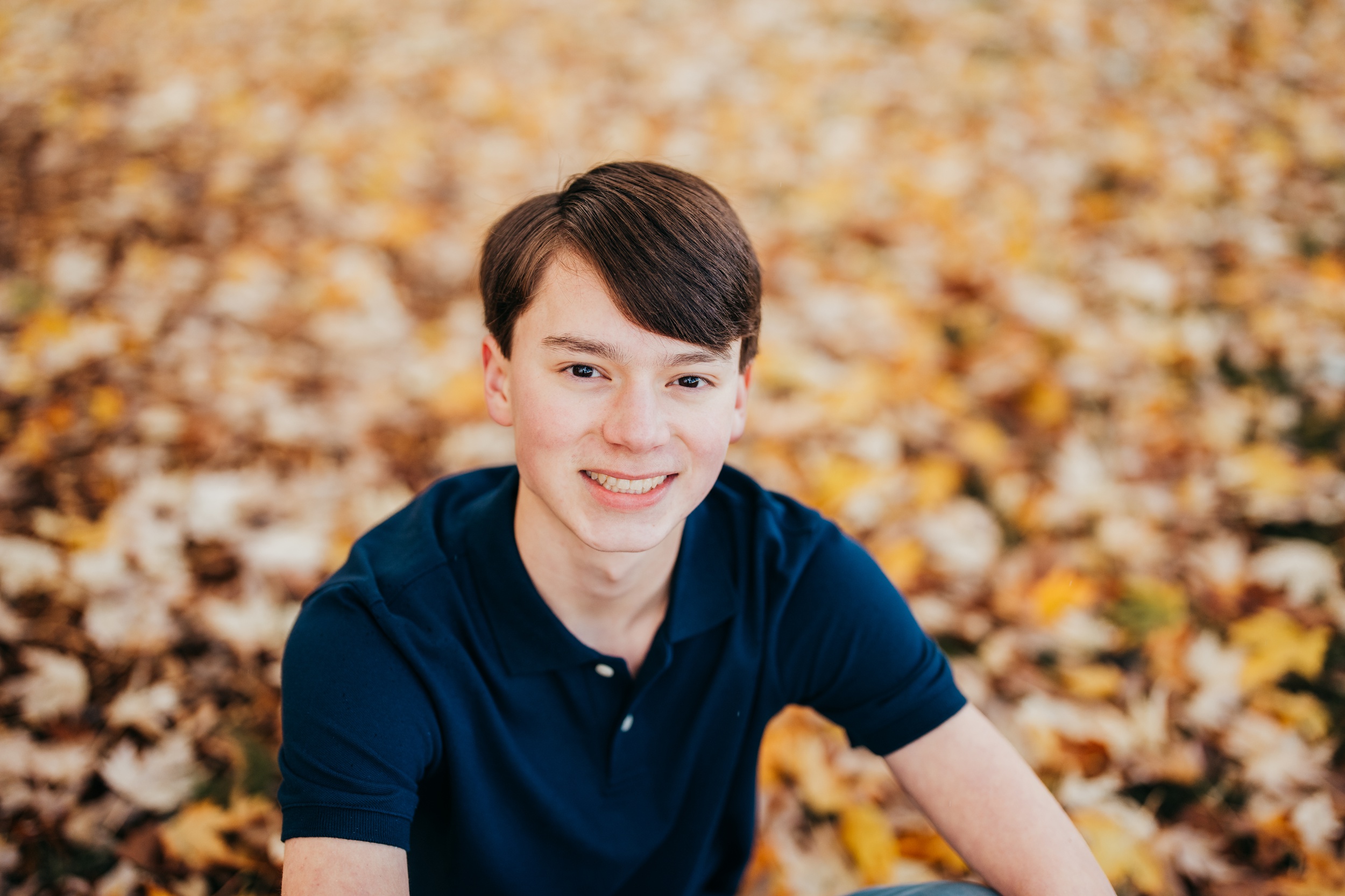 A happy high school senior in a blue polo sits in a lawn of fallen leaves smiling