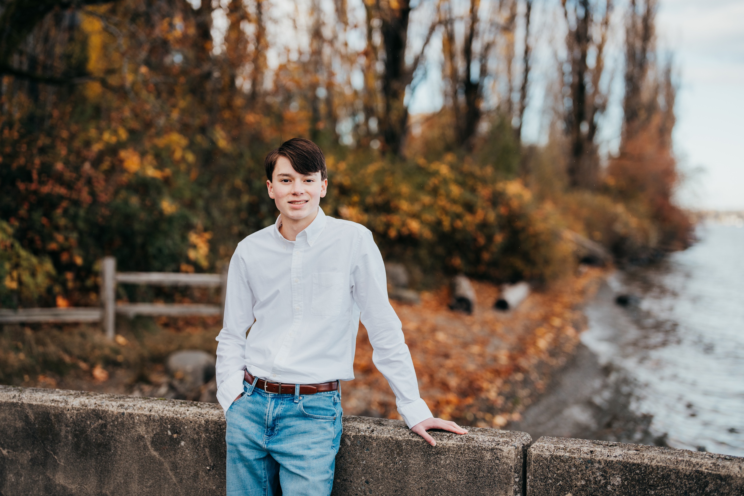 A teenage boy in a white shirt and jeans leans on a concrete wall by the water in fall after some act prep in seattle