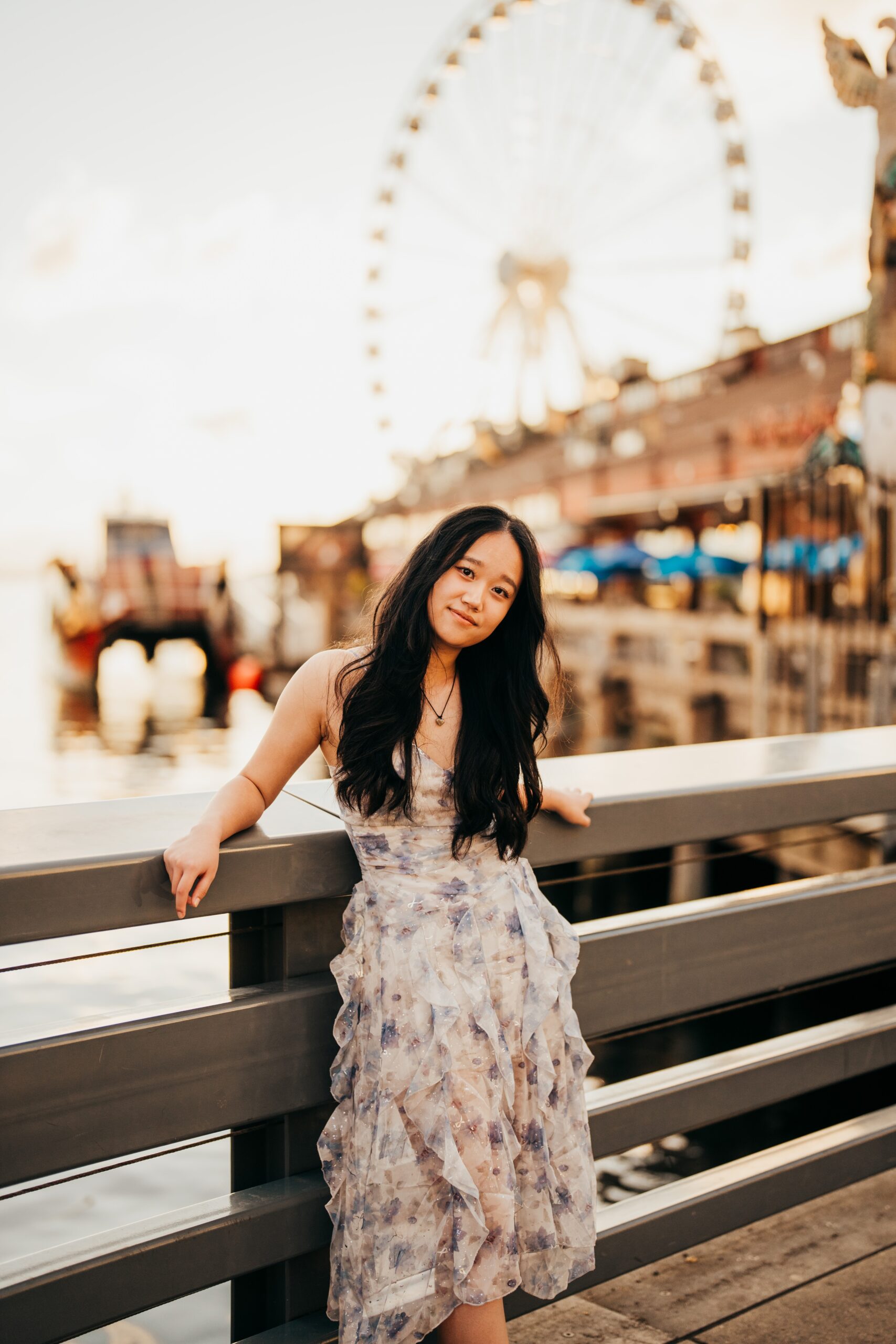 A high school senior in a floral dress leans on a dock railing in the wharf