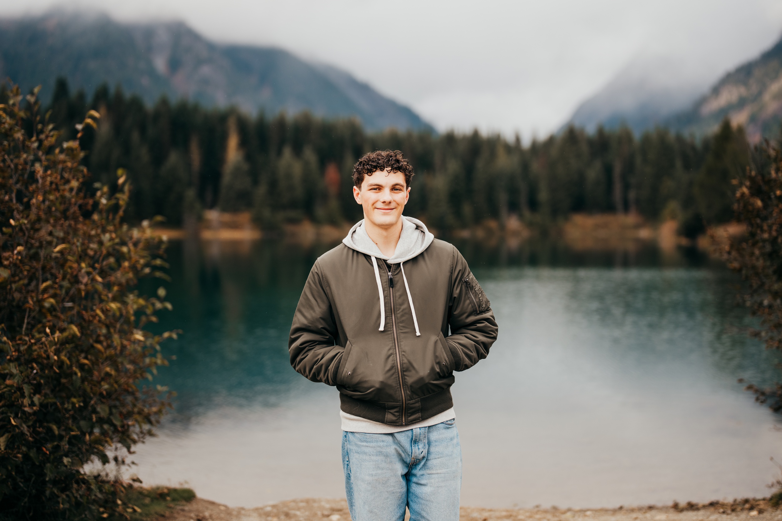A happy high school senior in a black jacket stands with hands in pockets on the edge of a lake
