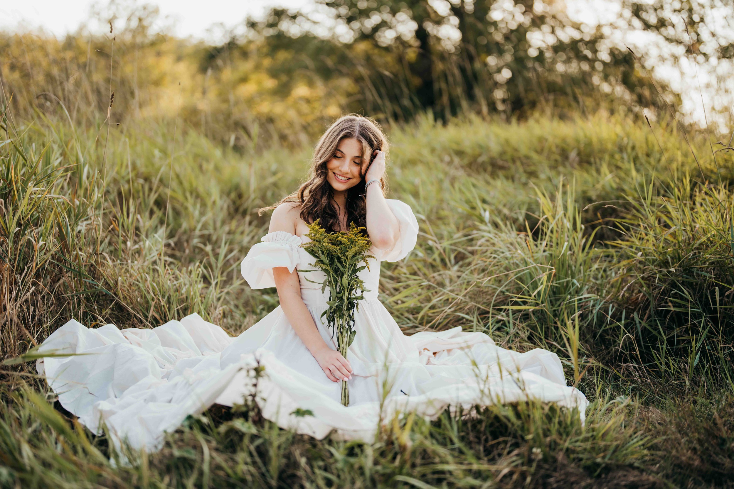A happy teenage girl sits in a field of tall grass at sunset holding some wildflowers in a white dress