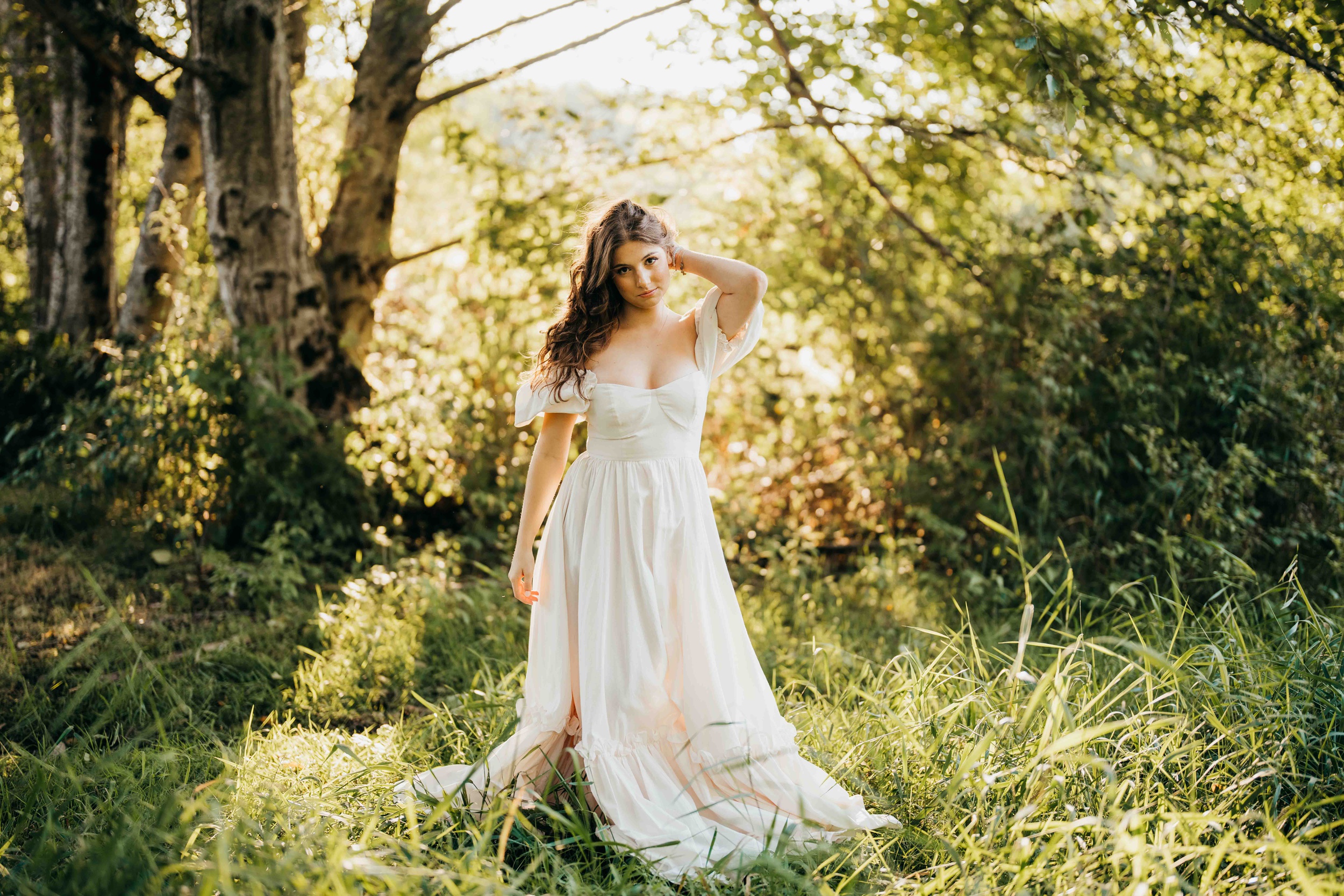 A teenage girl in a white dress pulls her hair back while standing in tall grass in a forest after visiting hair salons on mercer island