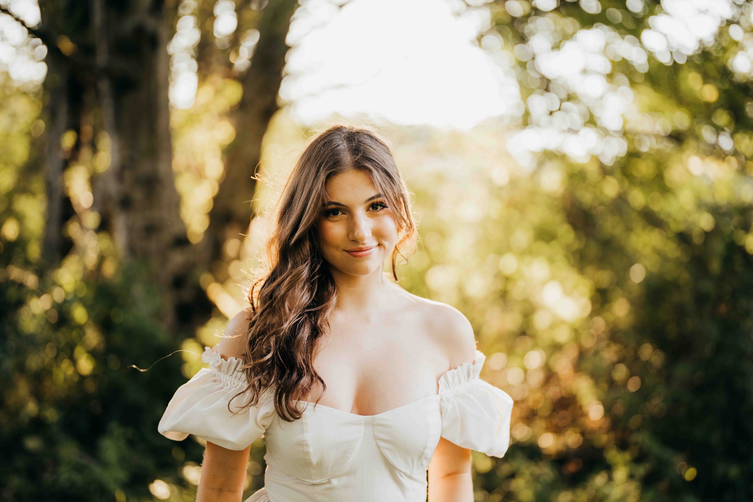 A high school senior with brown hair stands in a park at sunset in a white dress after visiting hair salons on mercer island
