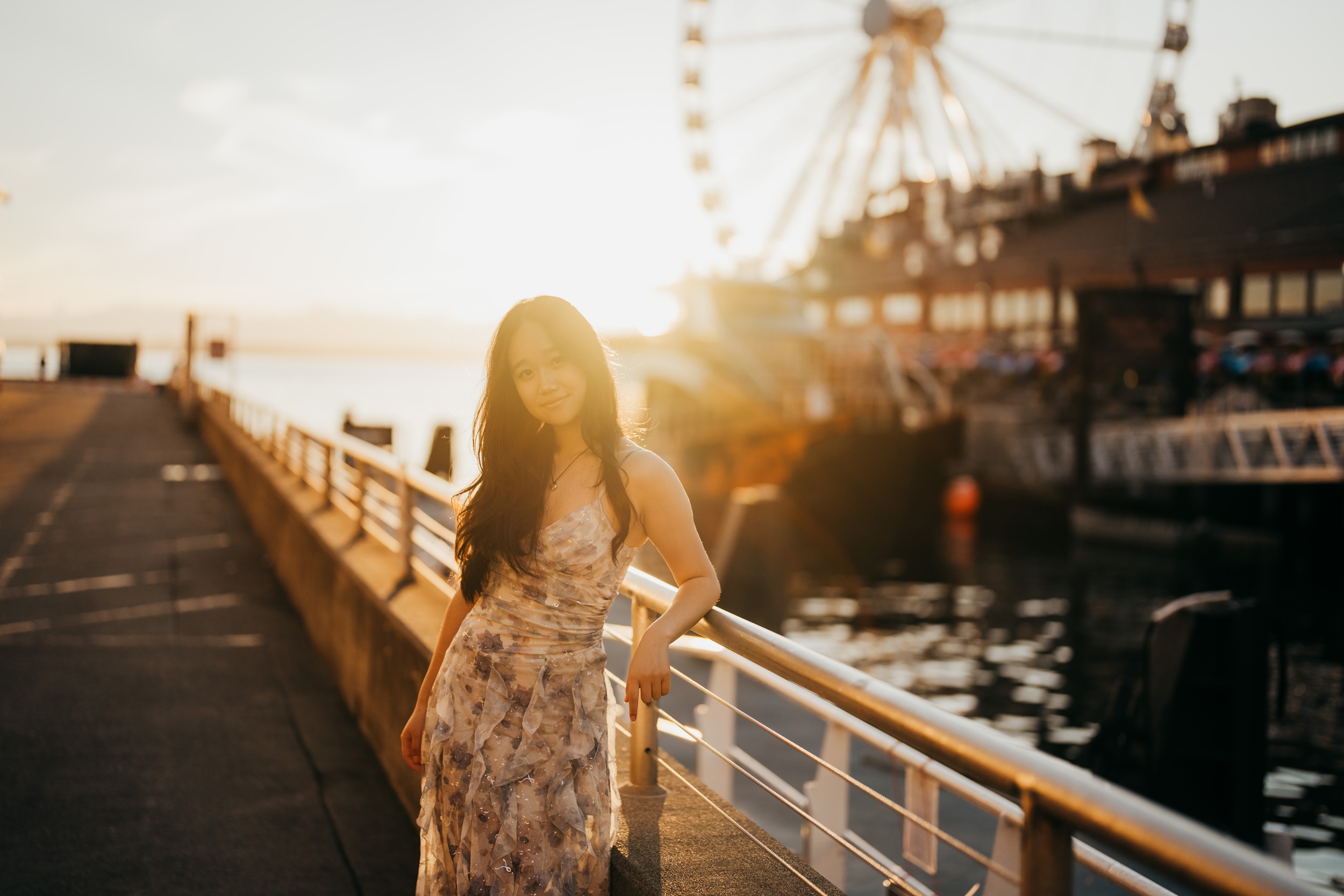 A high school senior leans on a dock railing by the fish market and ferris wheel at sunset