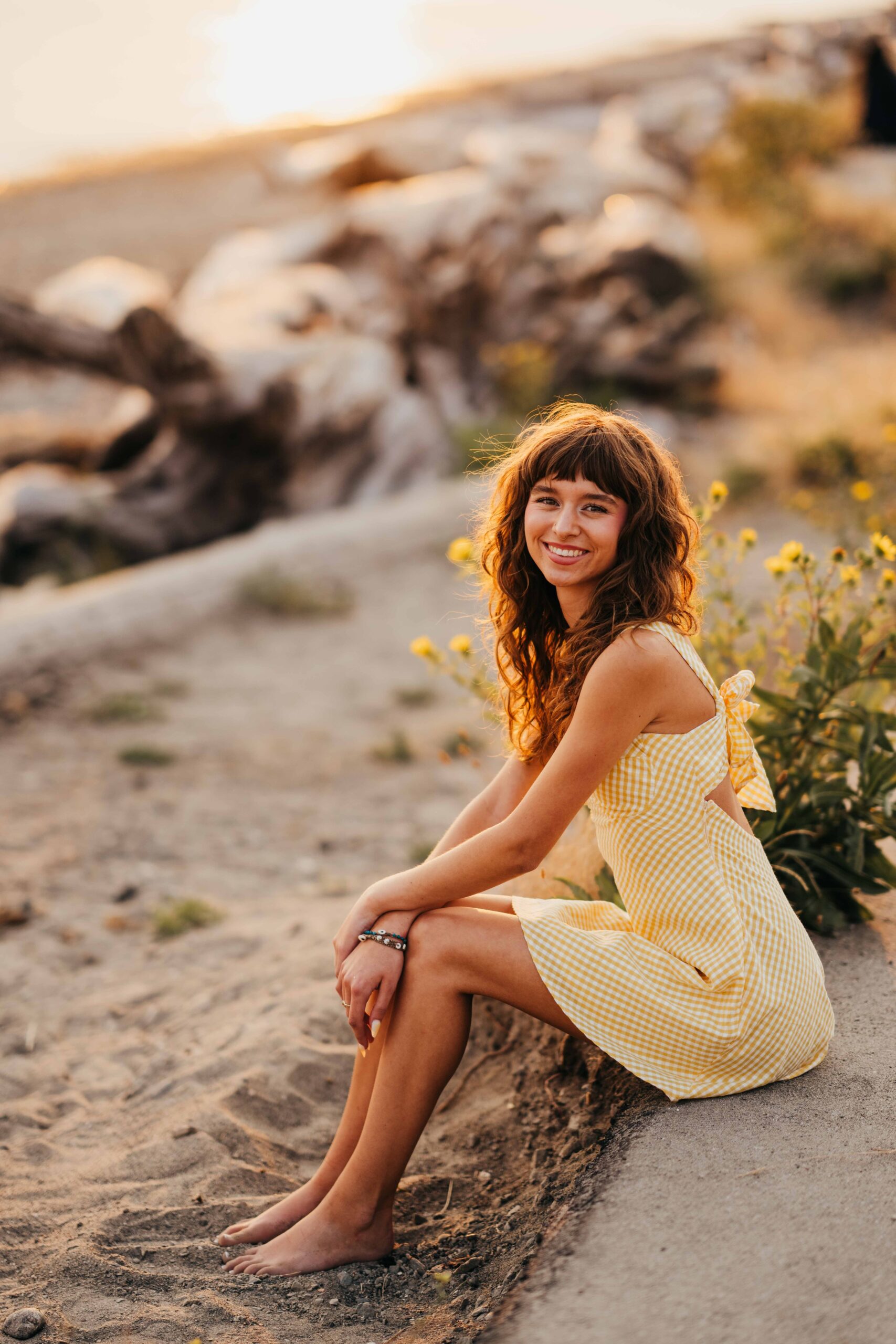 A happy high school senior sits on the edge of a beach in a yellow dress at sunset after shopping for homecoming dresses in seattle