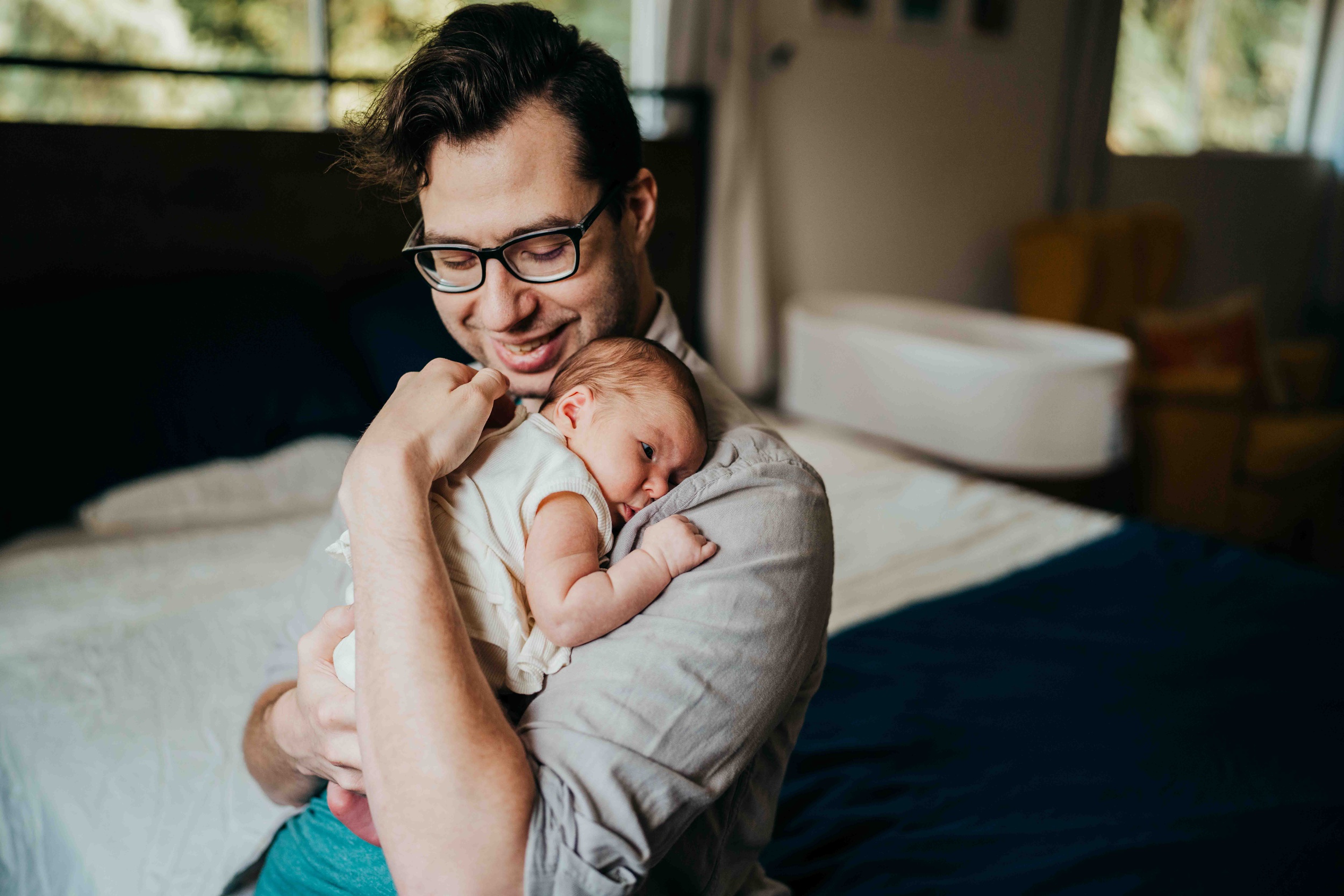 A happy new dad sits on a bed with his newborn laying on his shoulder