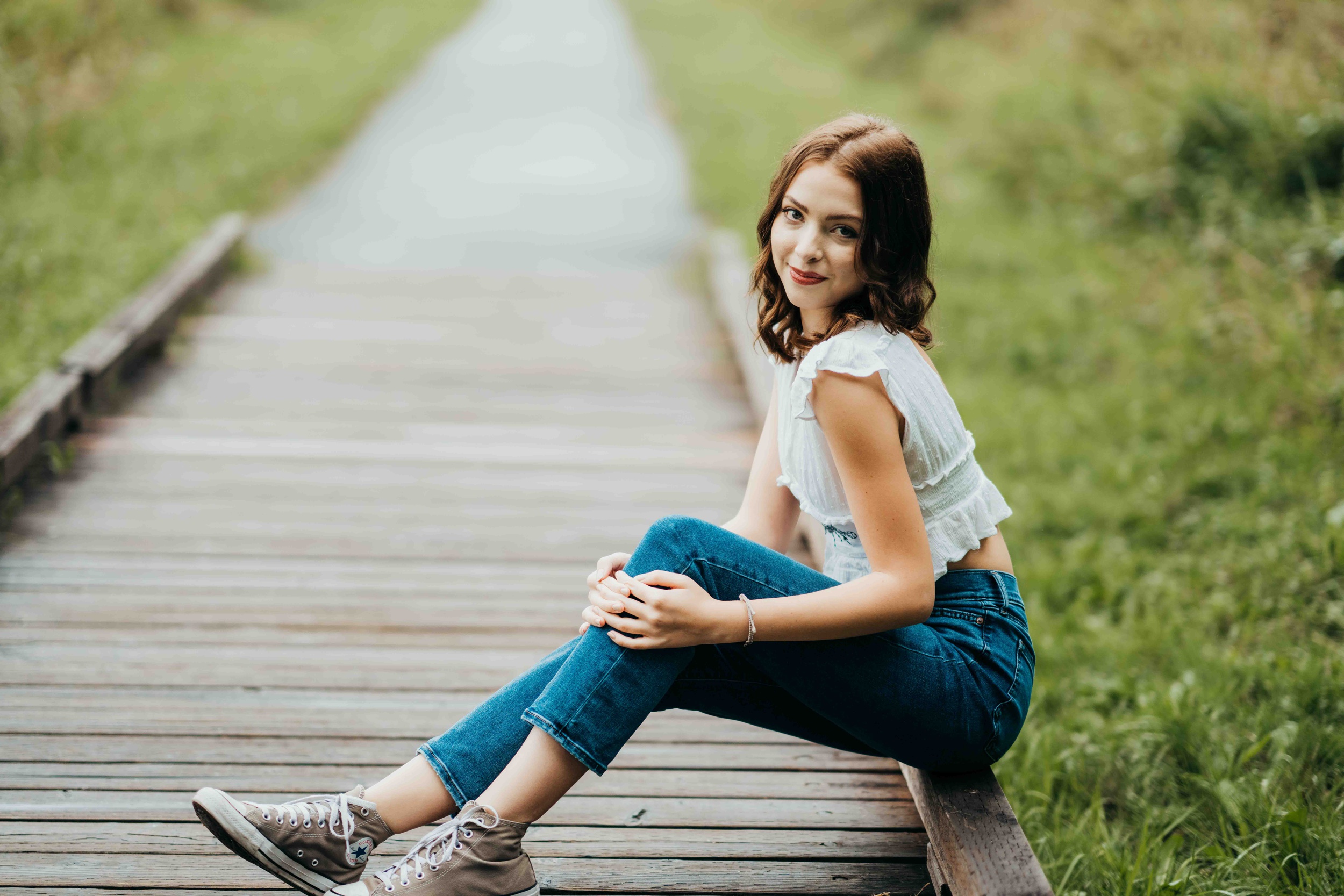 A teenage girl sits on the edge of a boardwalk in a park in a white blouse and jeans after using makeup artists on mercer island