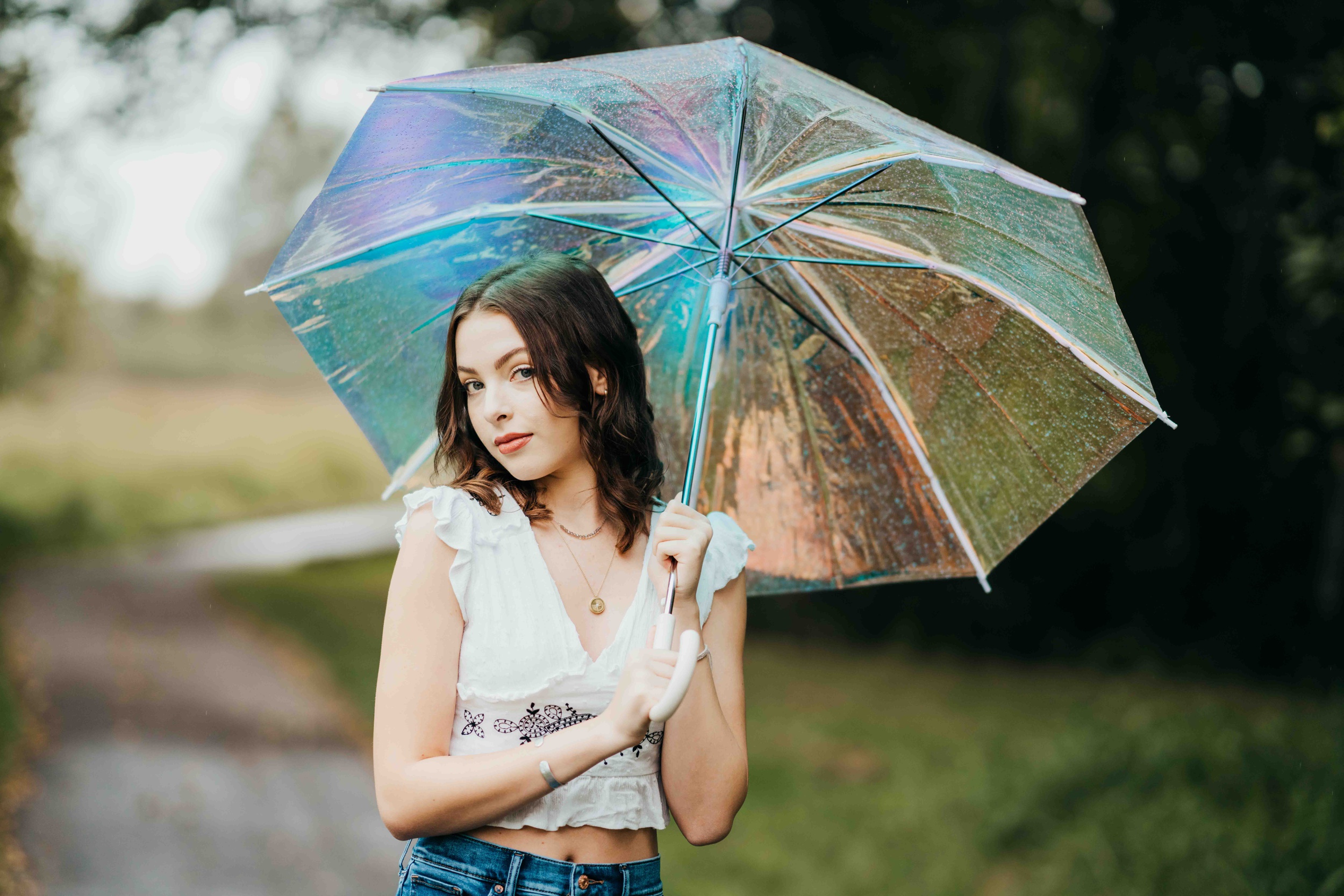 A teenage girl holds a clear umbrella in a white blouse and jeans in a park after finding makeup artists on mercer island