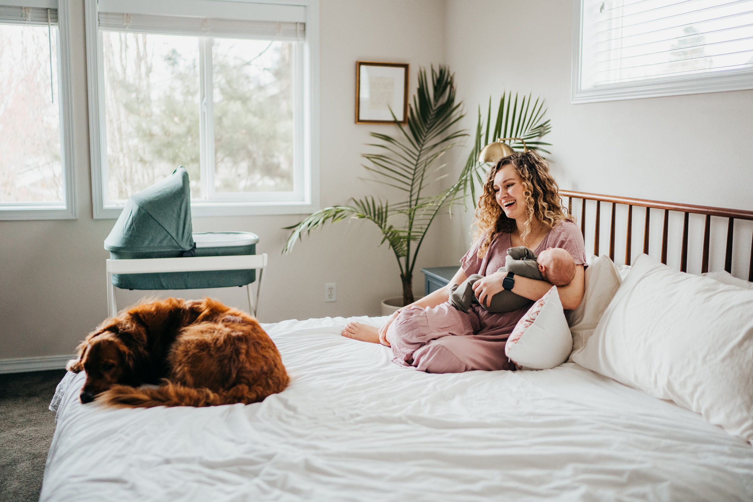 A new mom laugh while holding her newborn on a bed with their dog laying at the end of the bed