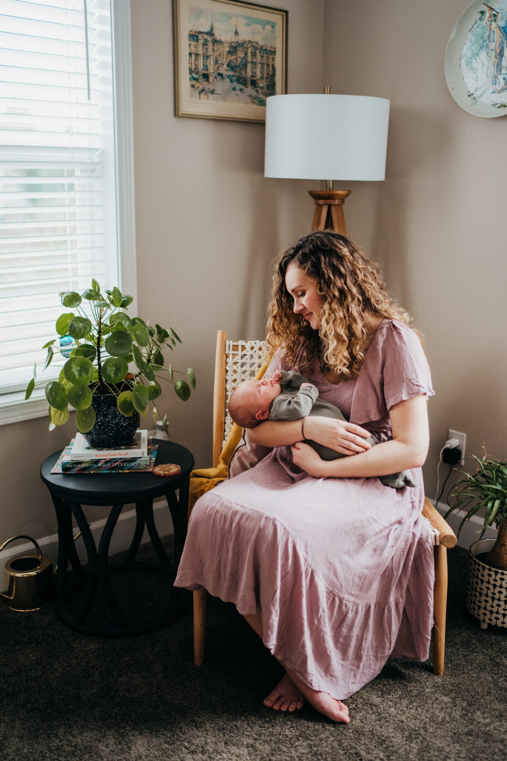 A happy new mom in a purple dress sits in a woven chair smiling down to her newborn sleeping in her arms after visiting mercer island pediatrics