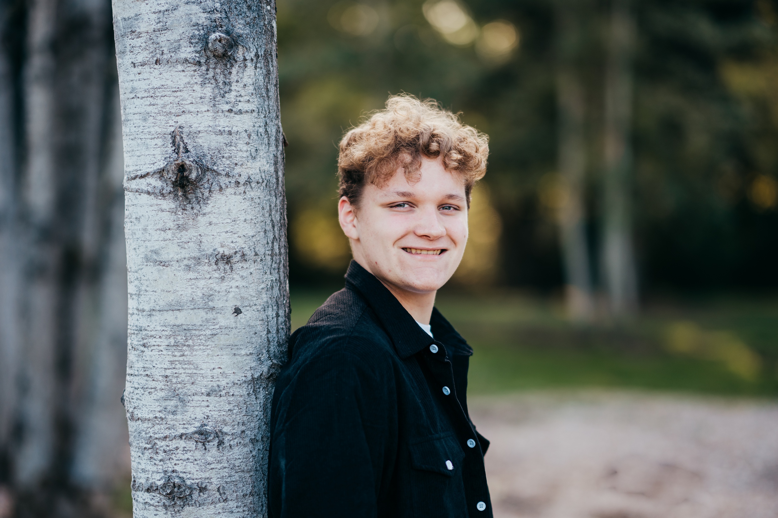 A happy high school senior leans on a tree in a black shirt while smiling