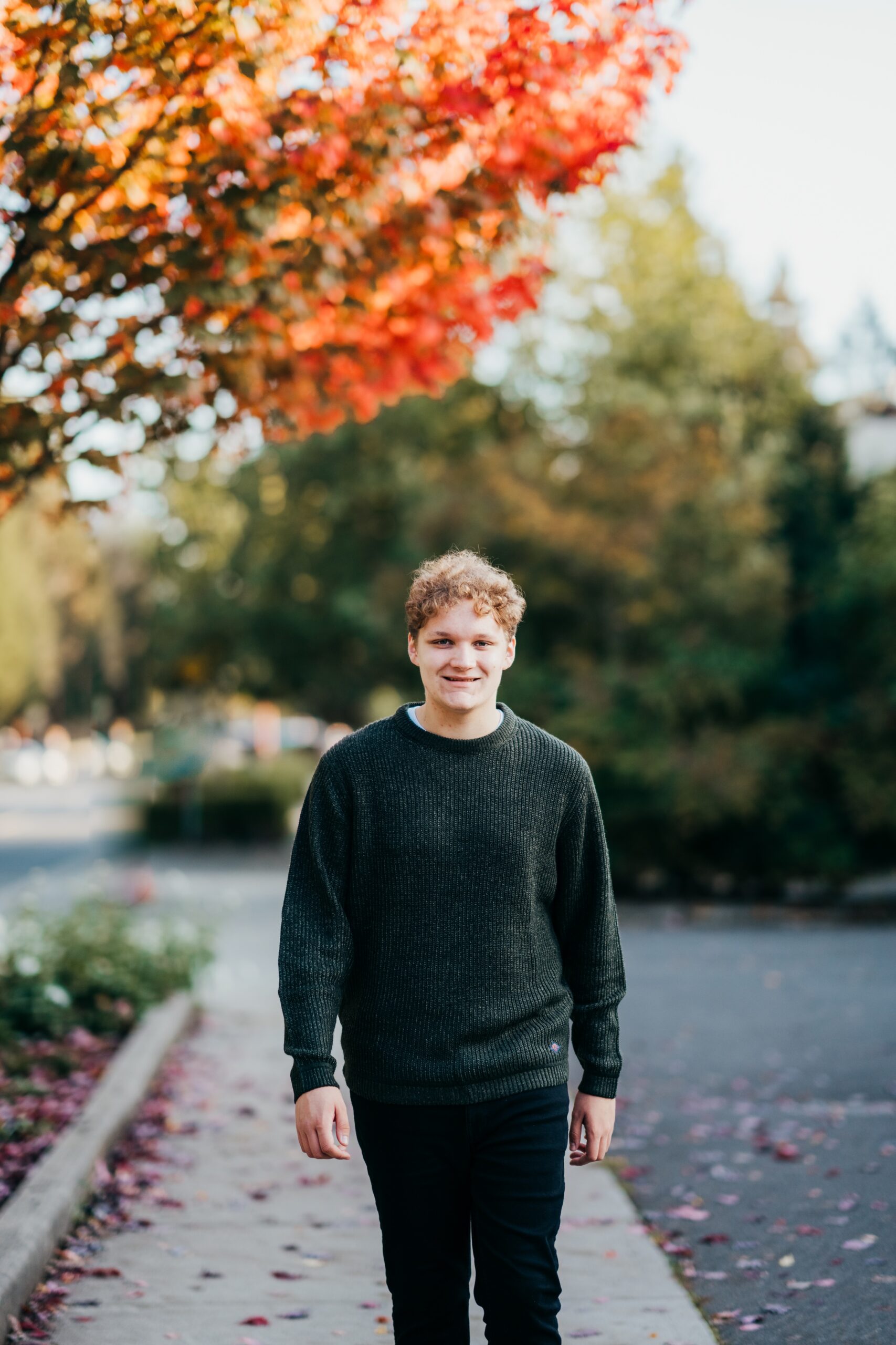 A high school senior in a green sweater and black pants walks under a red tree in fall smiling after some mercer island tutoring