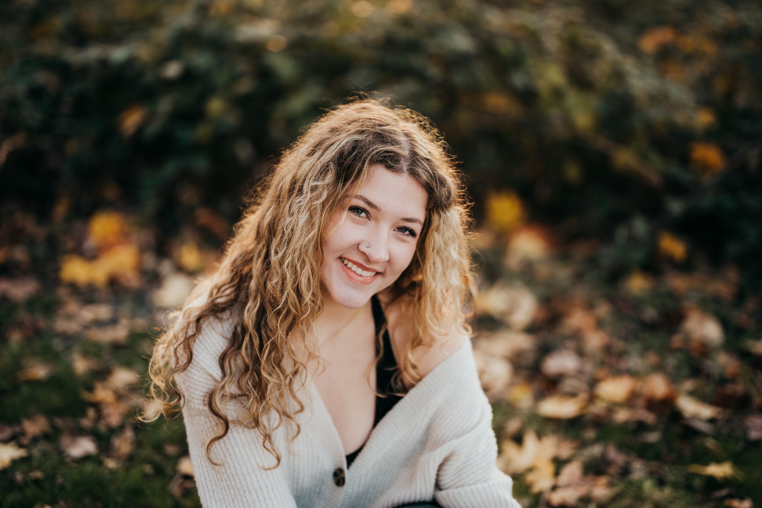 A smiling high school senior in a white sweater sits in a park lawn at sunset after visiting nail salons on mercer island