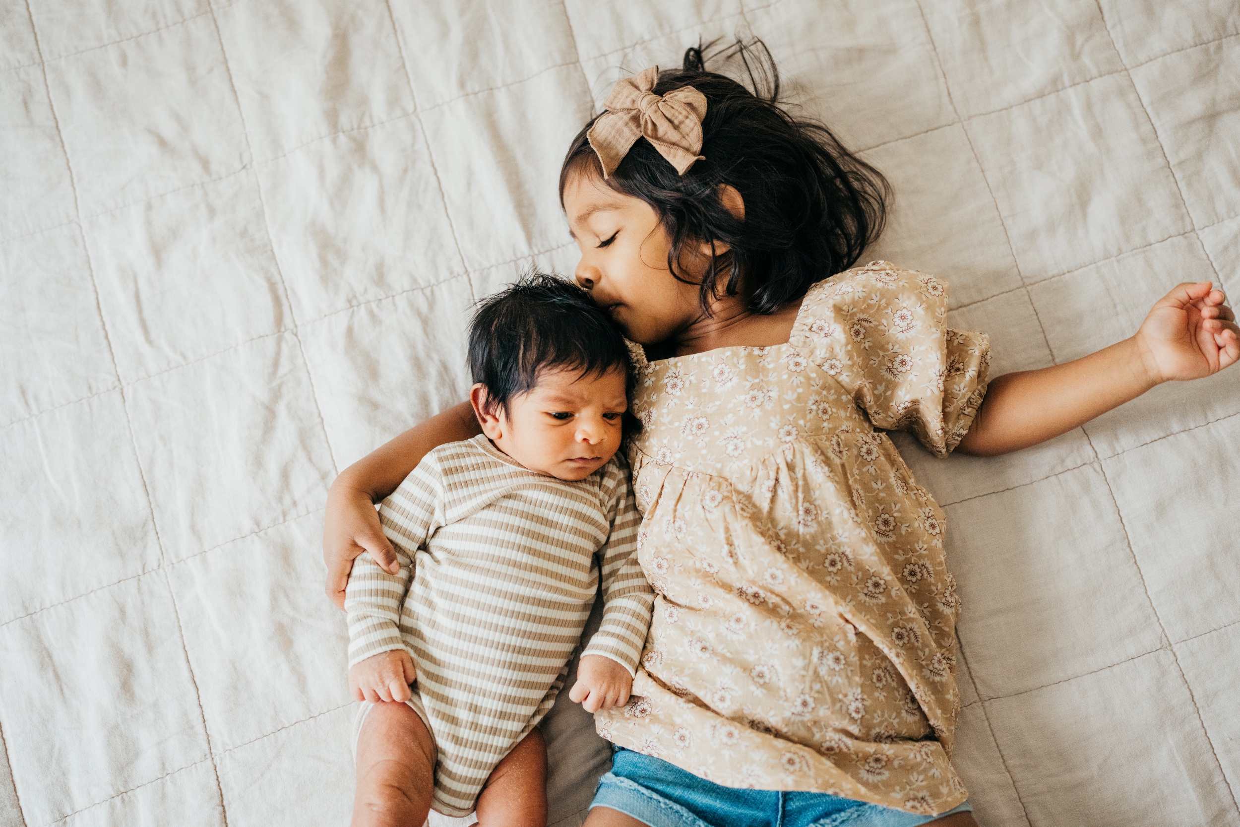 A toddler girl in a floral shirt kisses her newborn sibling while snuggling on a bed after meeting nannies in seattle