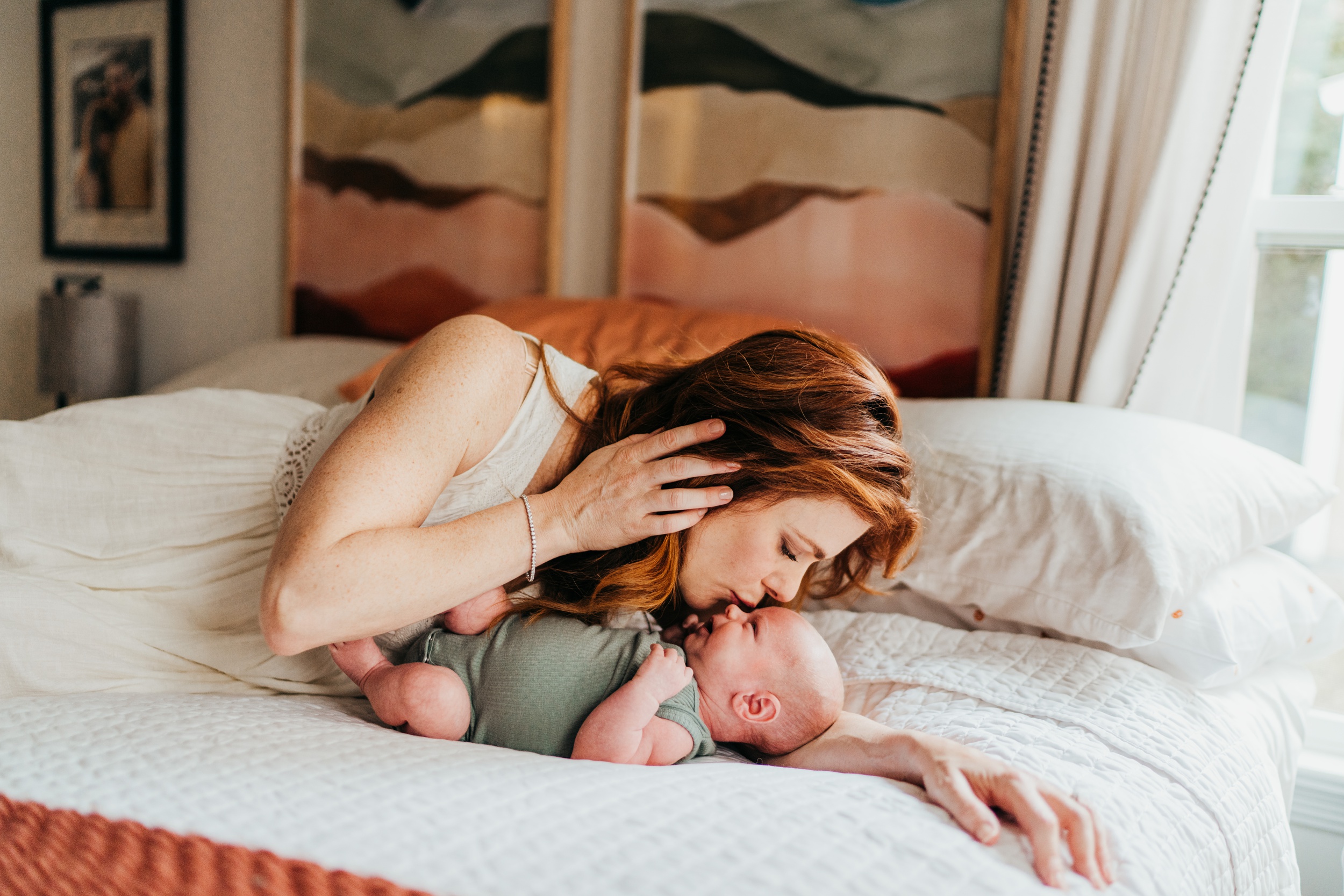 A new mom in a white dress lays on a bed kissing her newborn baby in a green onesie