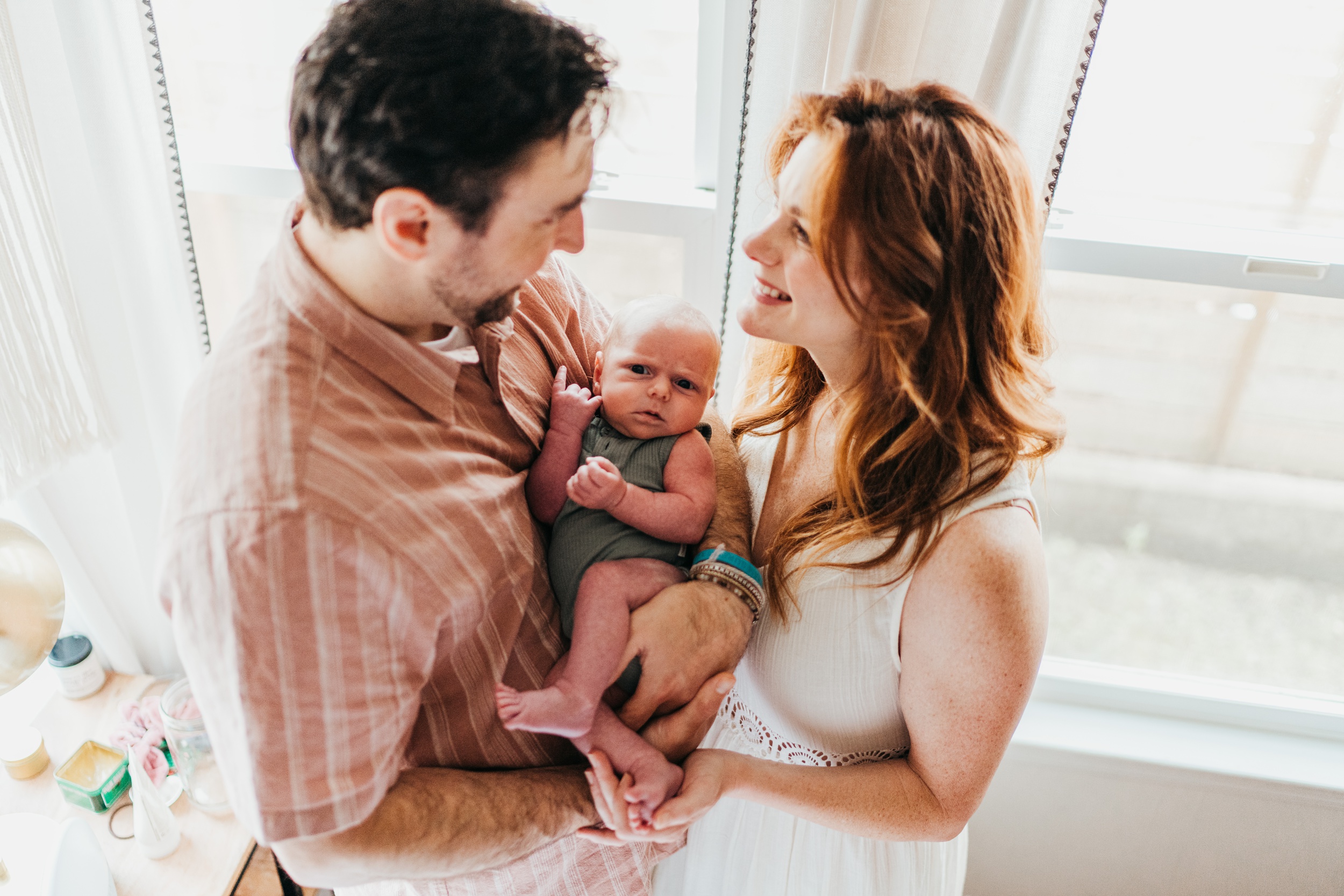 Happy new parents cradle their awake newborn in a window while smiling at each other after using northshore midwives