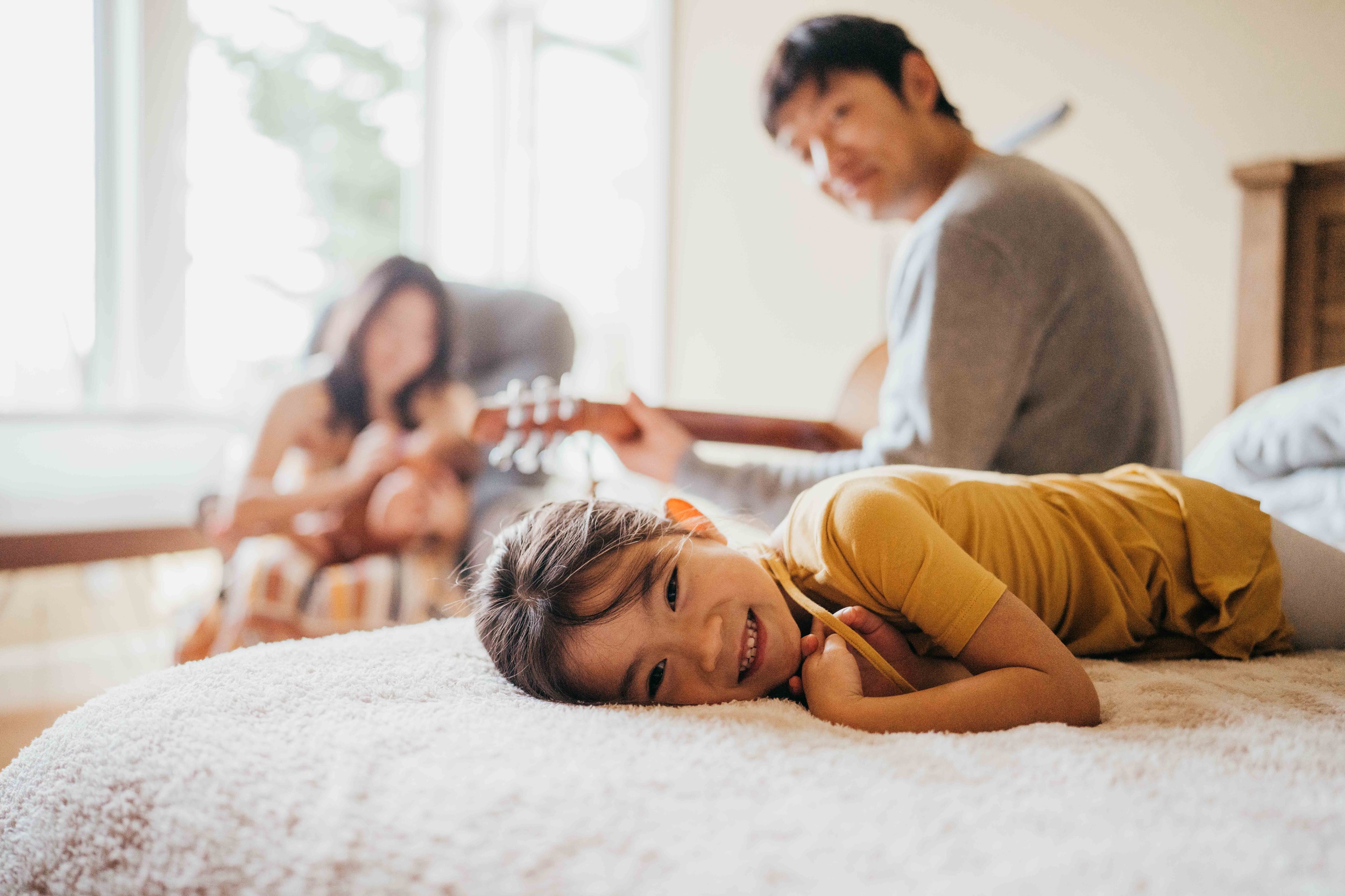 A young girl smiles while laying on a bed as dad plays guitar behind her