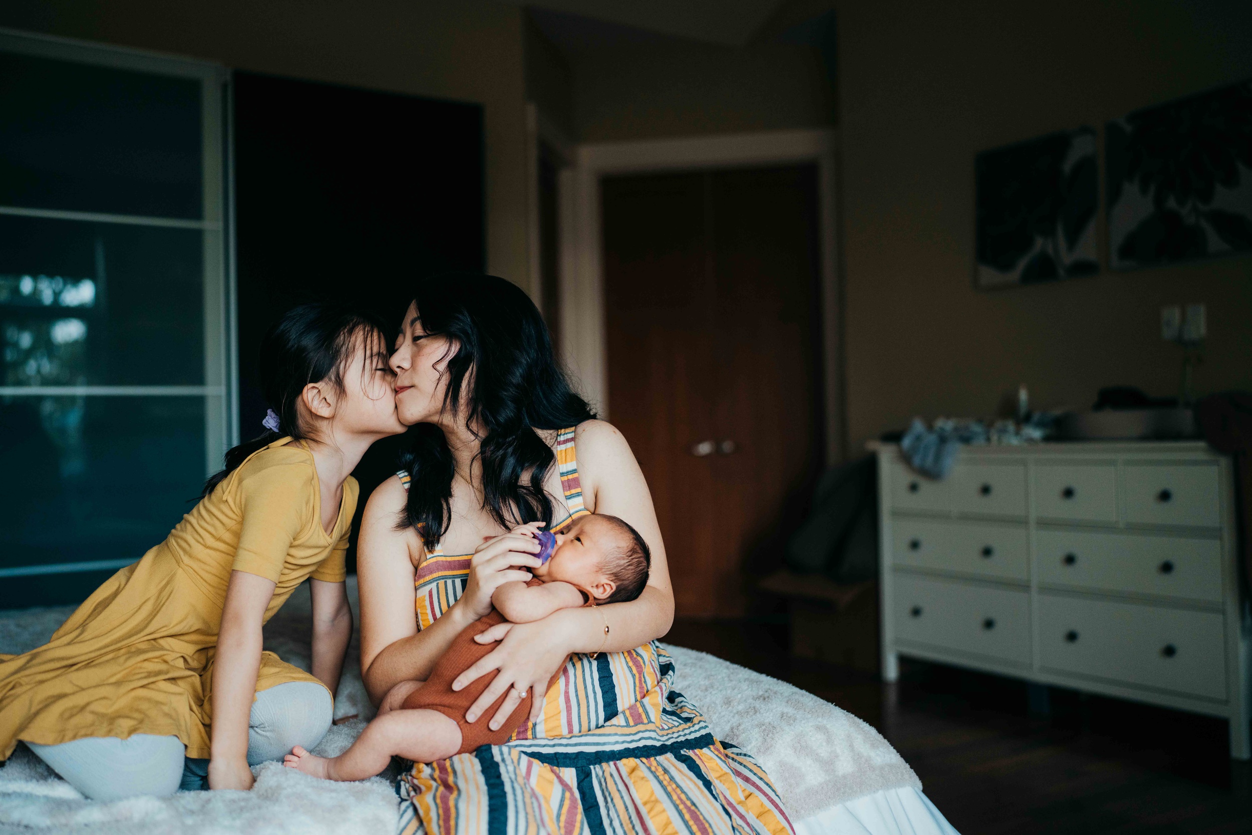 A young girl in a yellow dress kneels on a bed to kiss mom holding a newborn after visiting pediatricians in seattle