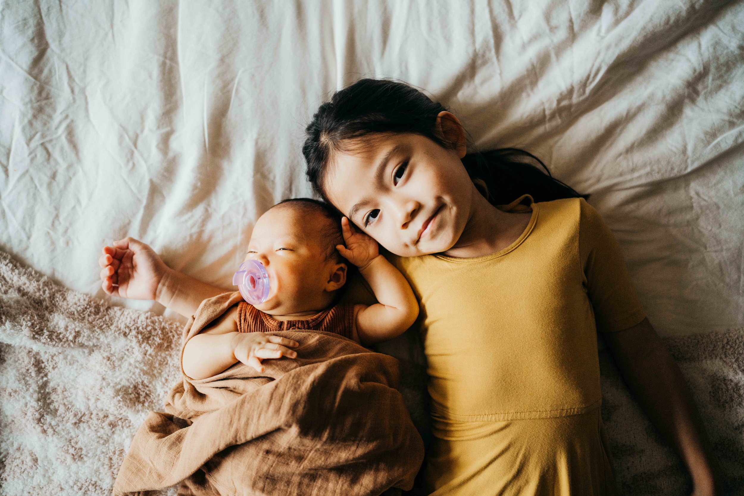 A toddler girl in a yellow dress cuddles her newborn sibling on a bed under a blanket after visiting pediatricians in seattle