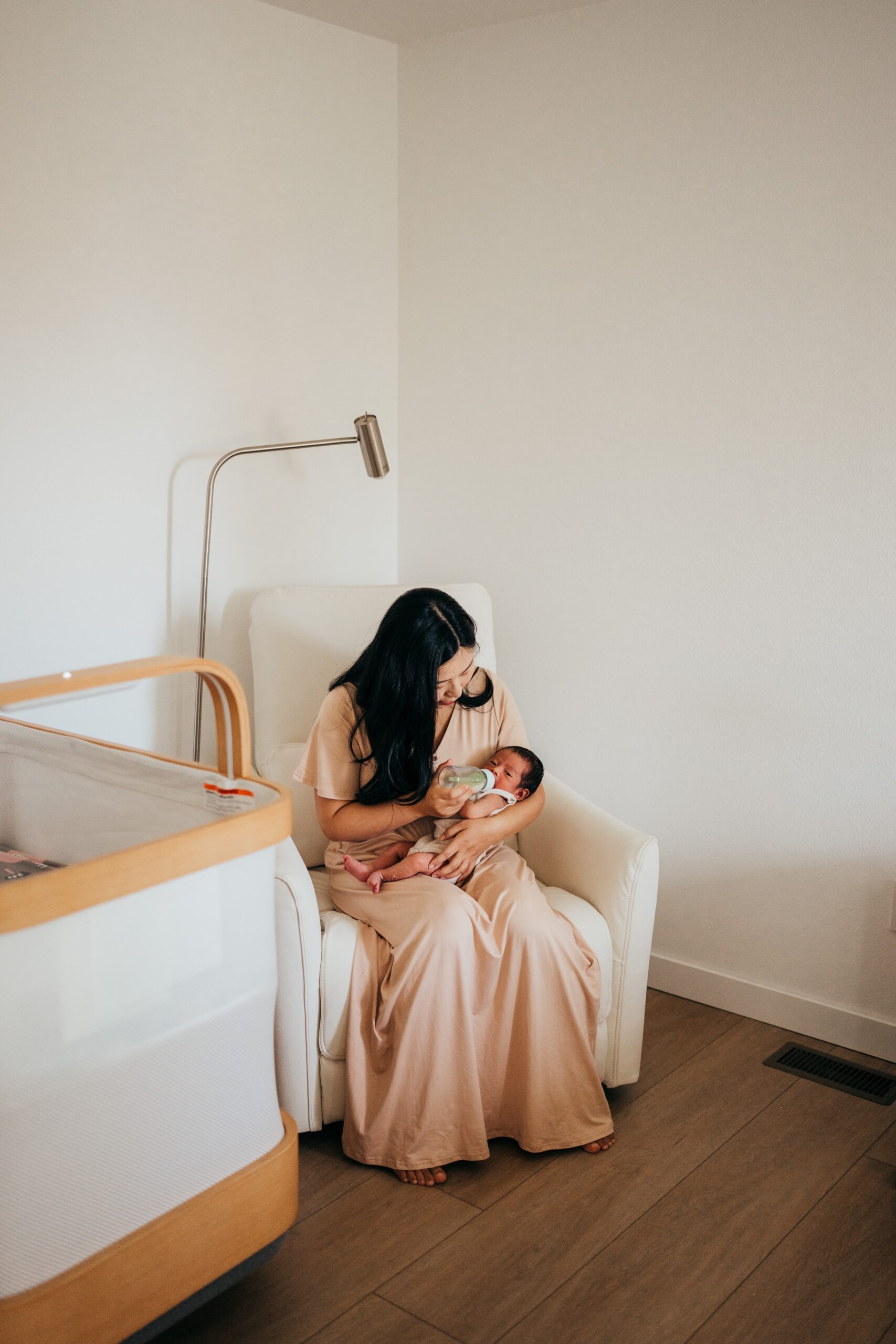 A mother in a cream dress sits in a nursing chair feeding her baby after meeting postpartum doulas in seattle