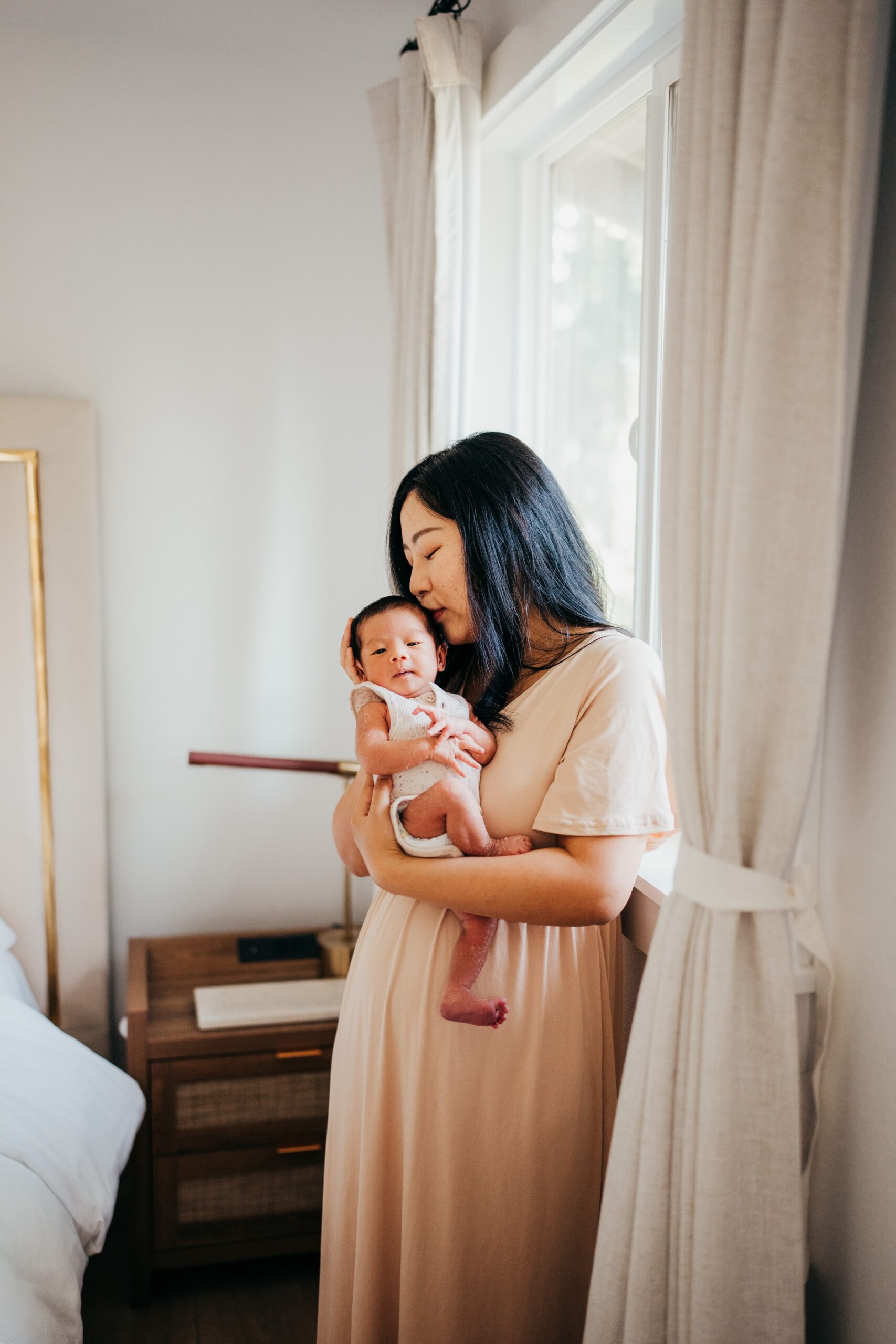 A new mom kisses her newborn baby in her arms while standing in a bedroom window thanks to postpartum doulas in seattle