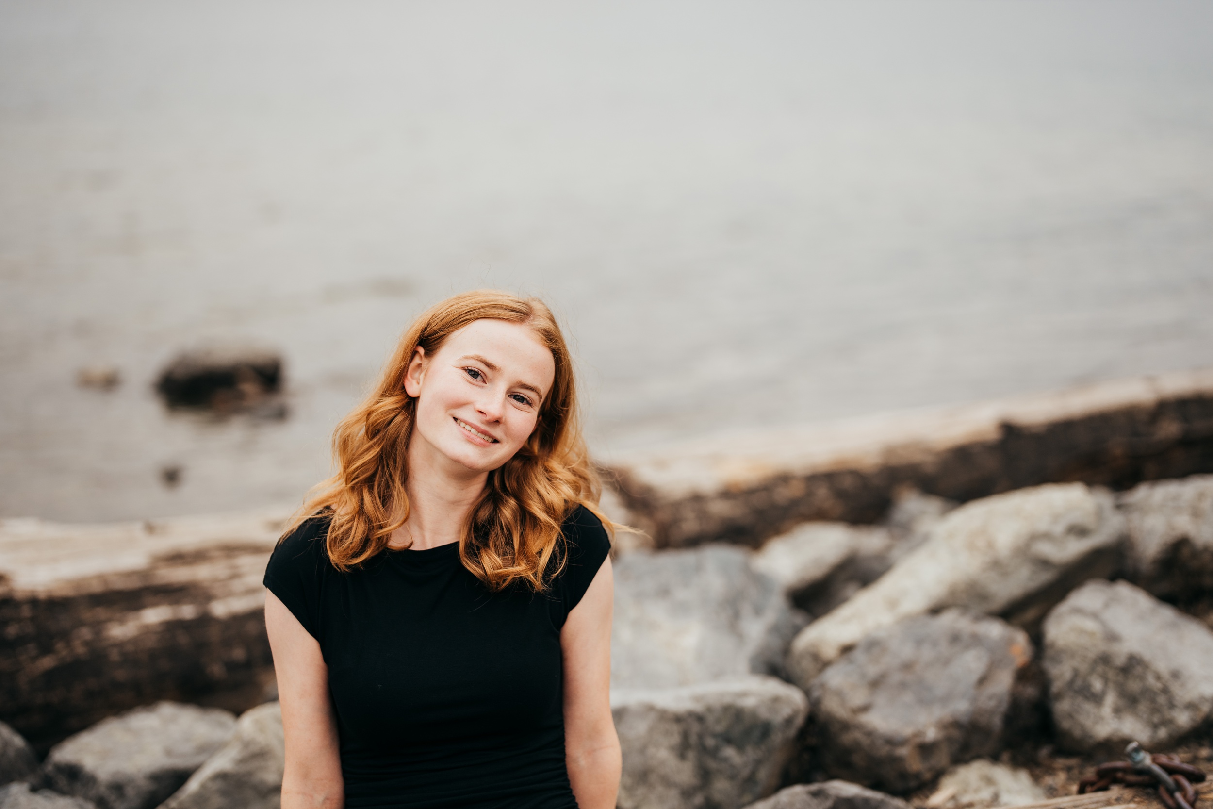 A happy high school senior stands on rocks by the water in a black shirt