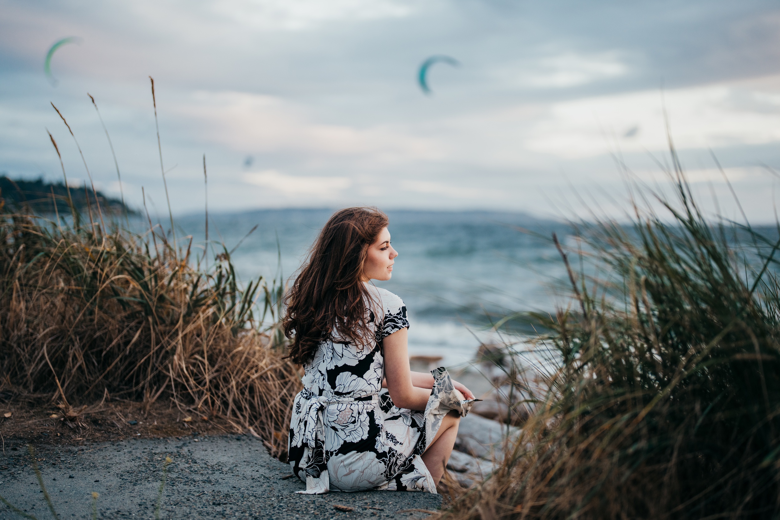 A woman in a floral dress sits on a beach dune watching the water