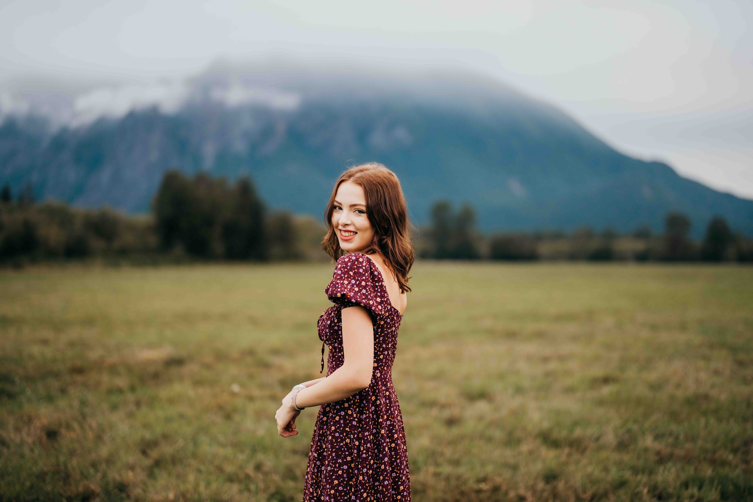 A happy high school senior walks in a field near a mountain in a purple dress while smiling over her shoulder after exploring prom dress shops in seattle