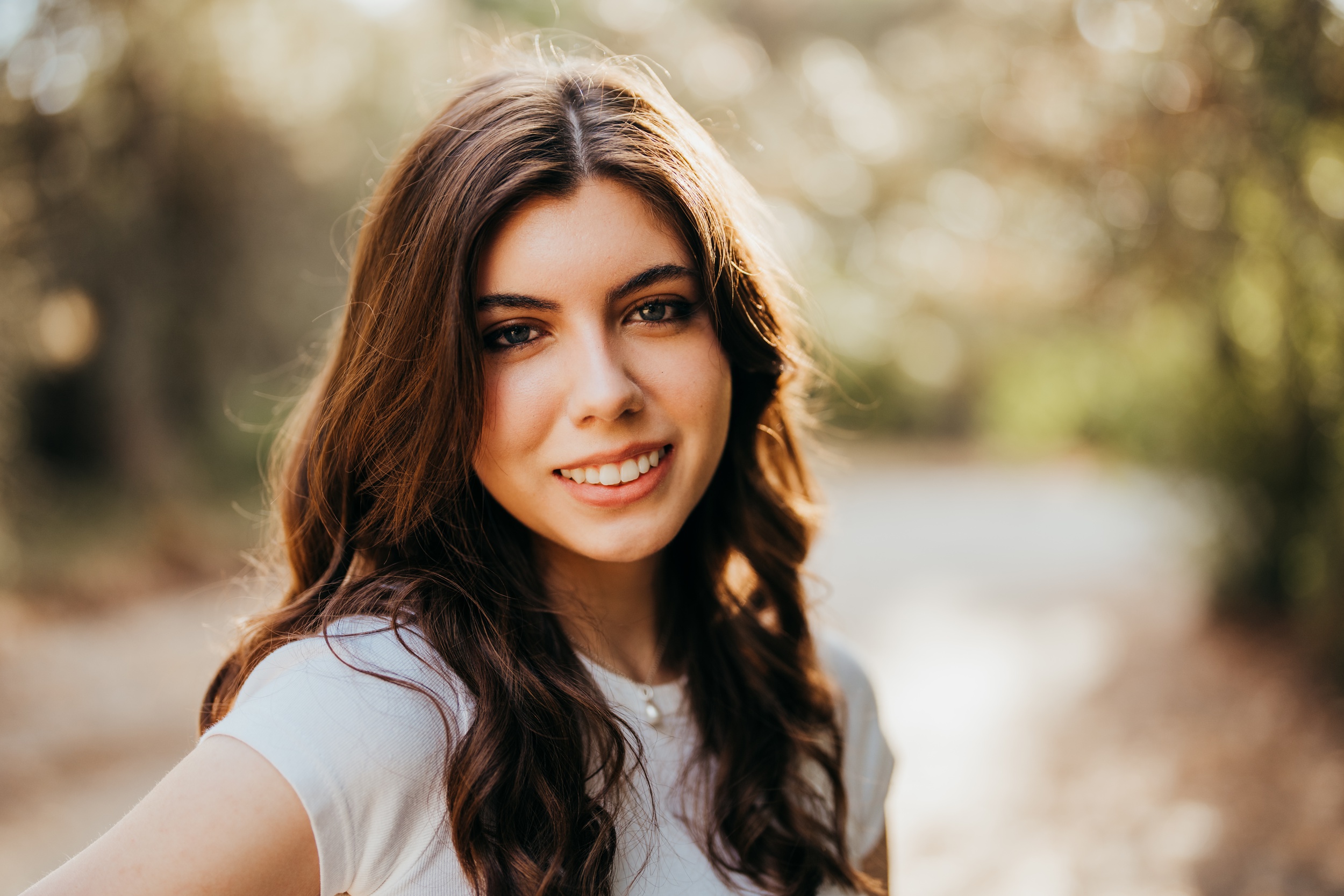 A smiling high school senior in a white top looks over her shoulder in a park