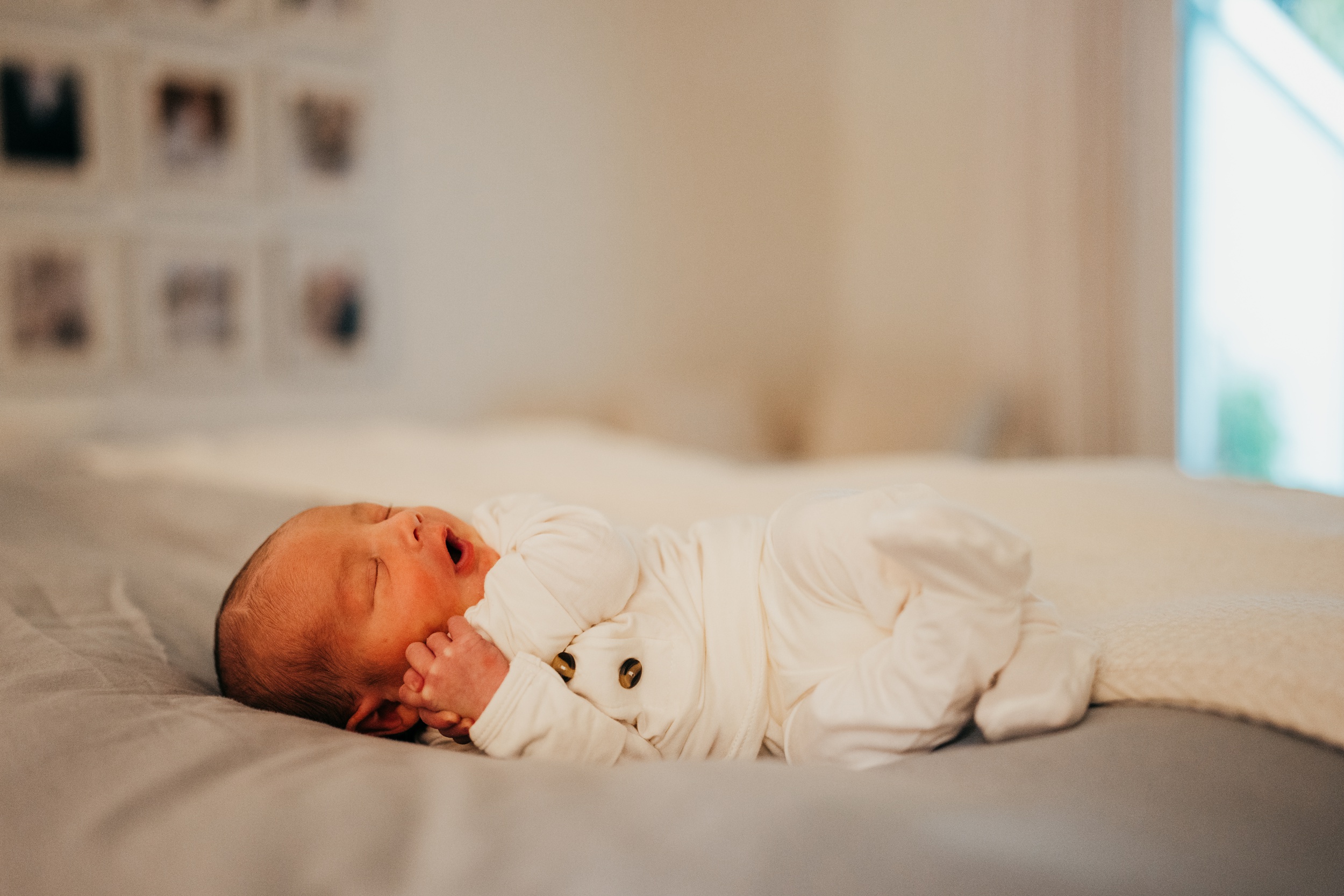 A newborn baby sleeps in a white onesie on a bed with mouth open