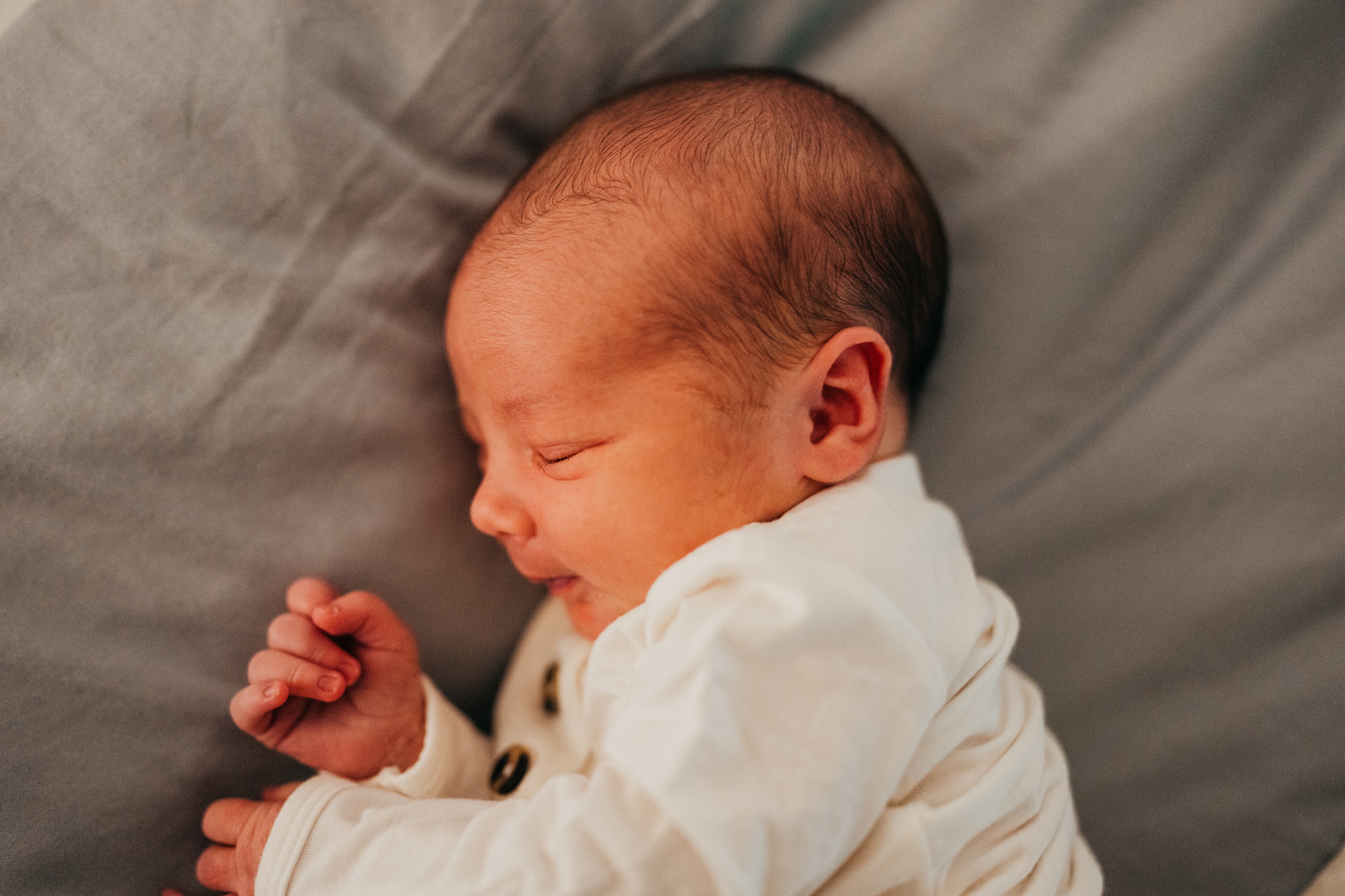 A newborn baby sleeps on a grey bed in a white onesie with a smile thanks to sleep training in seattle