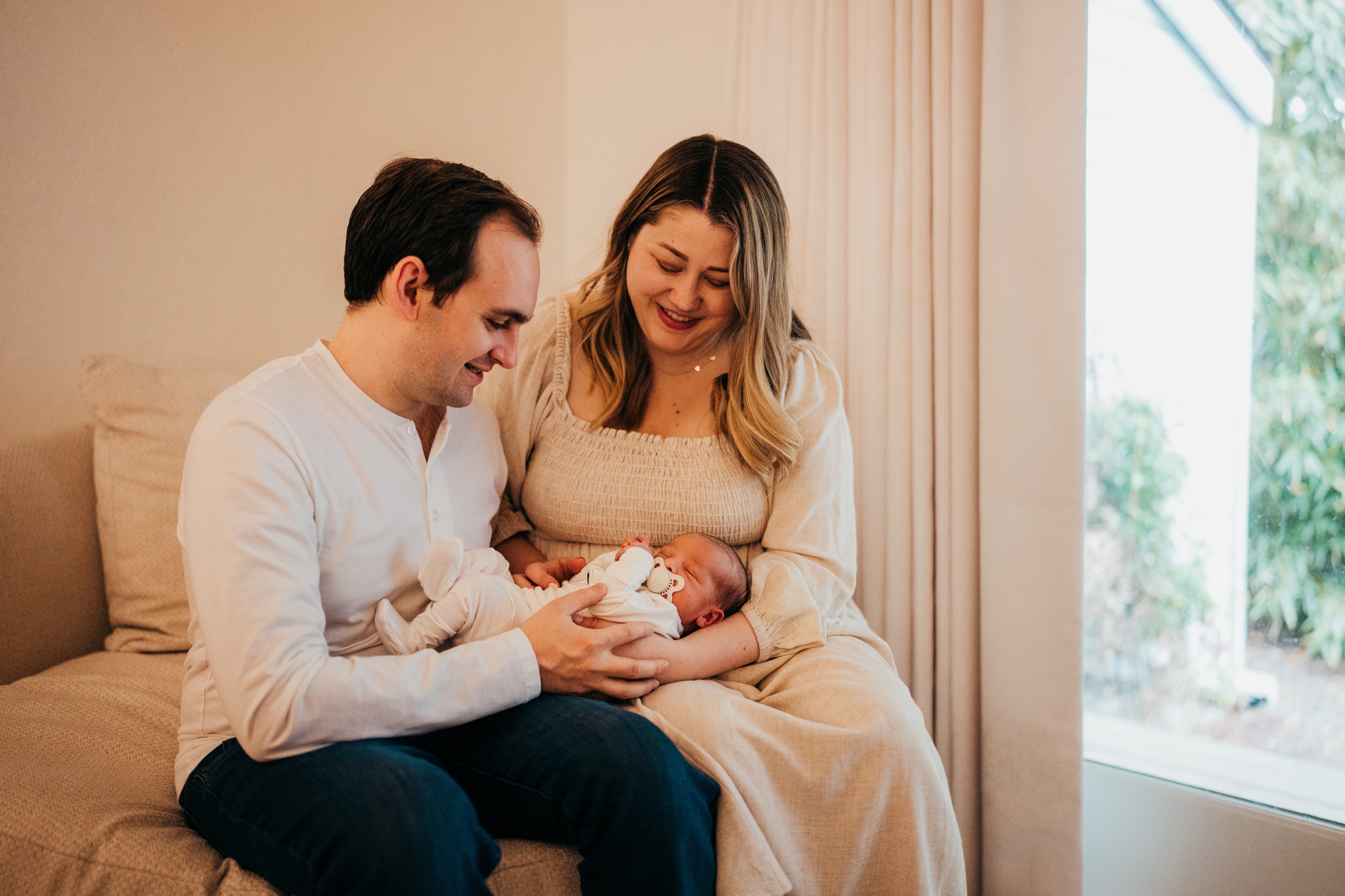 Happy new parents sit on a couch in a window smiling down at their sleeping newborn in their lap thanks to sleep training in seattle