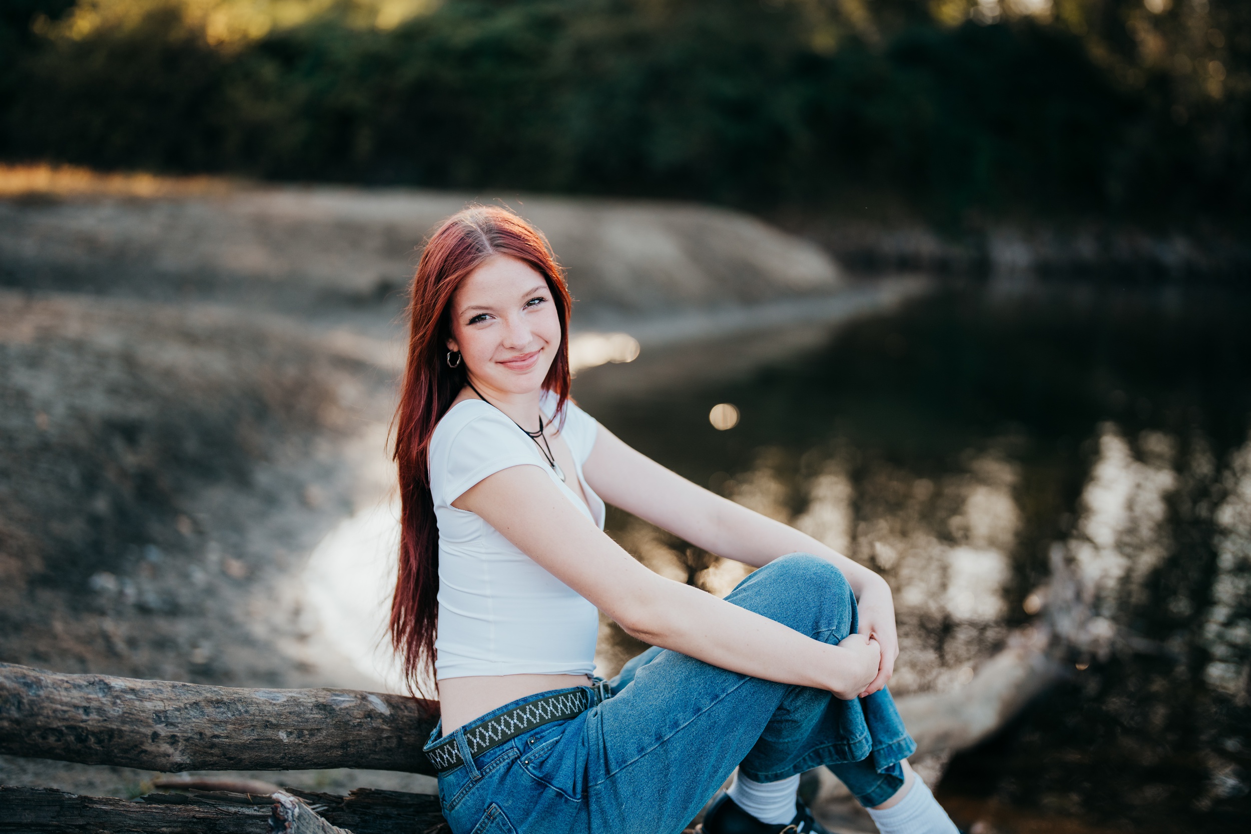 A high school senior in a white top and jeans sits on the edge of a pond at sunset smiling