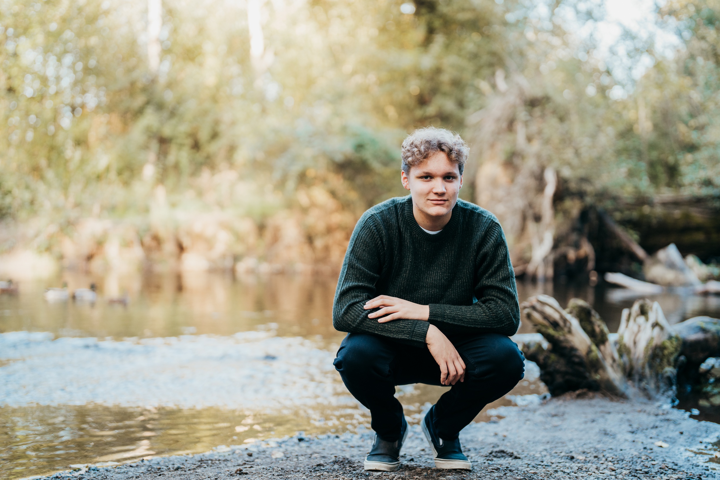 A high school senior squats by the water in a green sweater