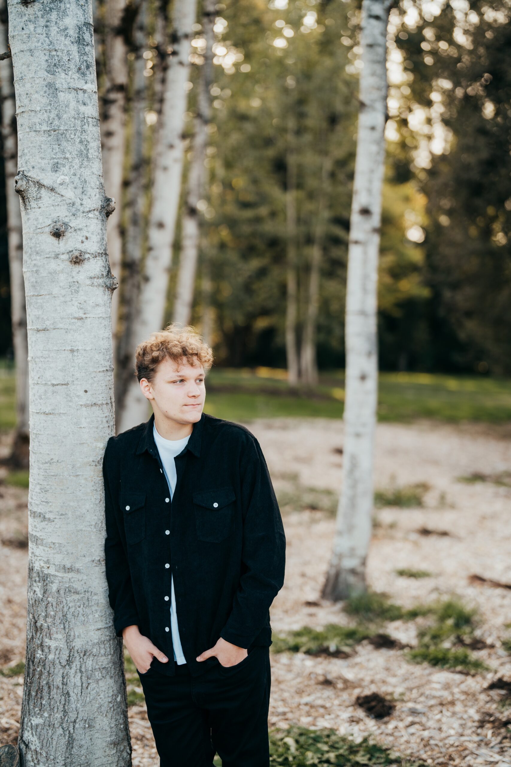 A high school senior in black leans on a tree in a park at sunset after finding tux rental in seattle