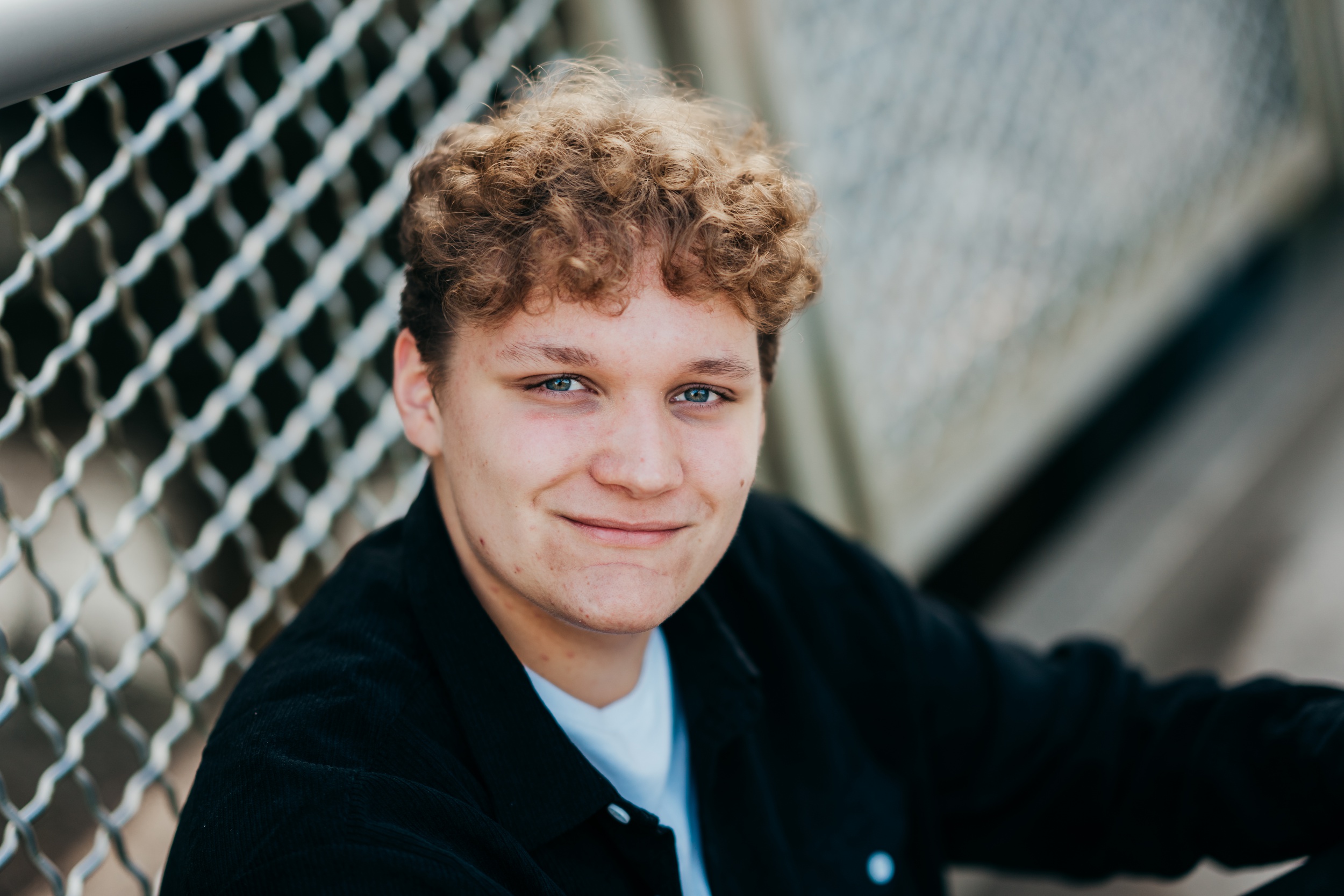 A high school senior sits against a chain link fence in a black shirt after finding tux rental in seattle