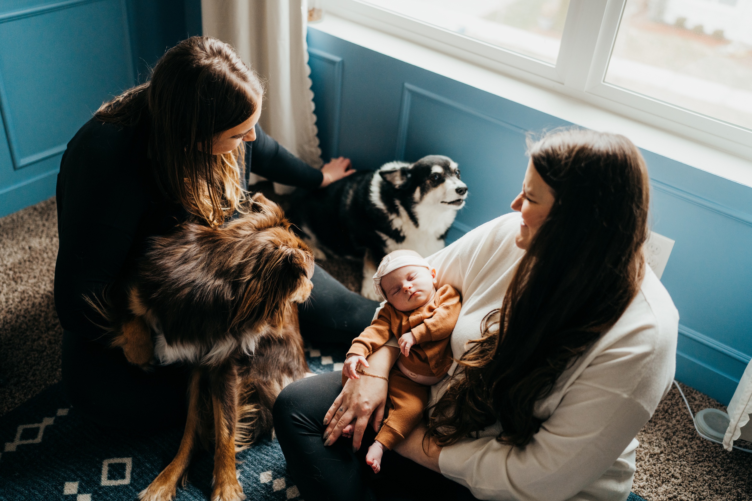 Happy new moms sit on the floor under a window with their two dogs and newborn sleeping before some baby music classes in seattle