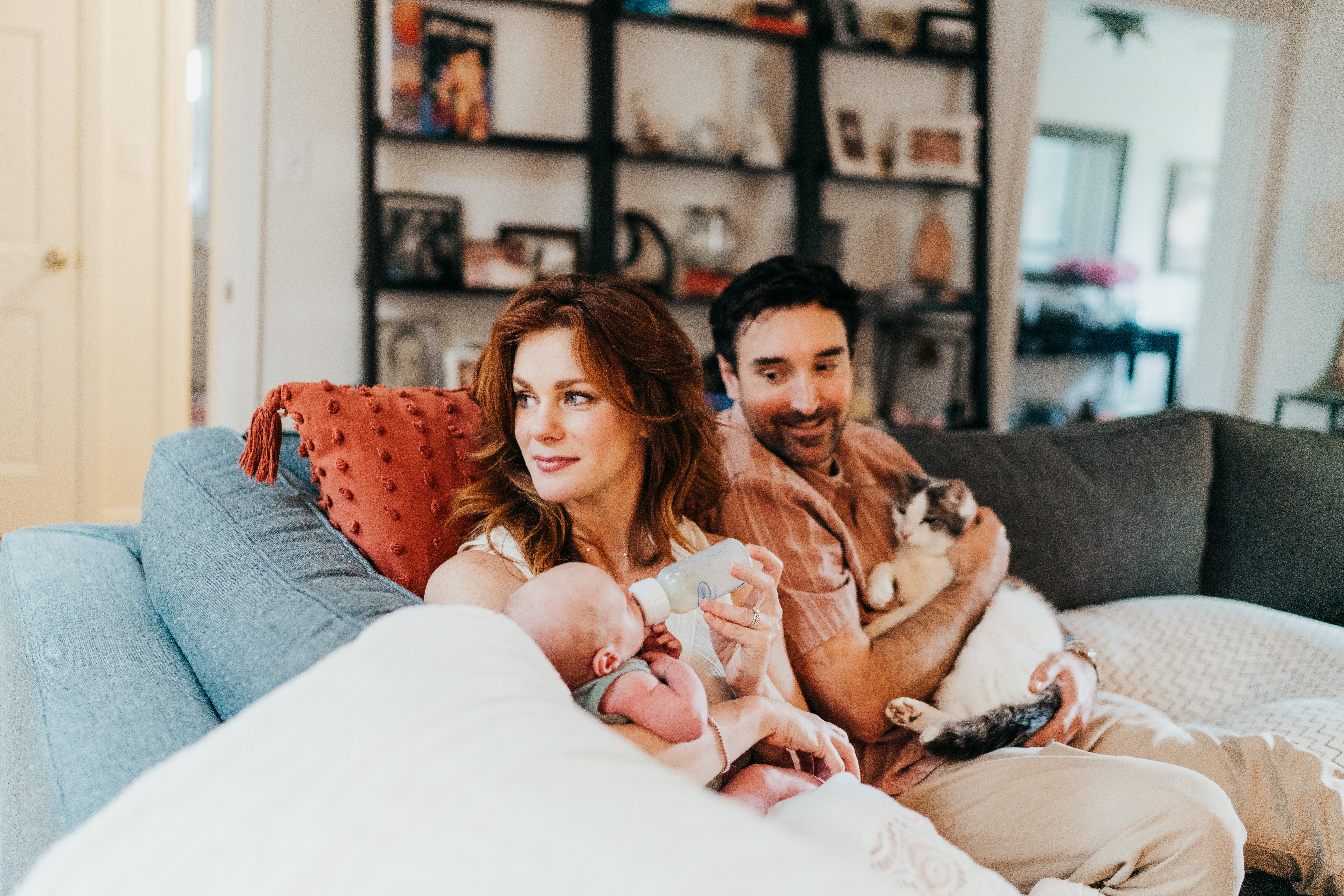 A new mom sits on a couch bottle feeding her newborn as dad snuggles a cat after finding baby proofing in seattle